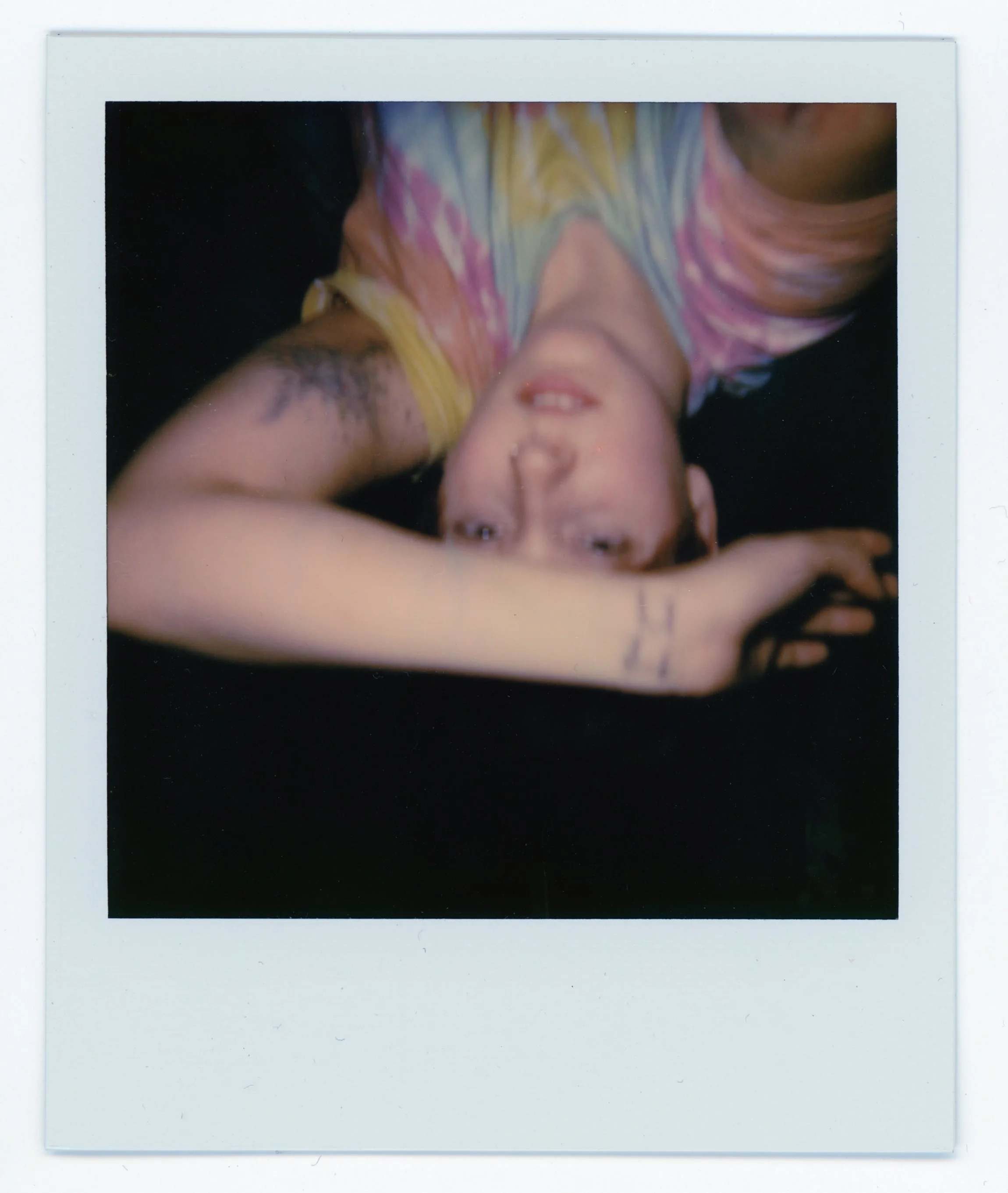 A person lying on their back taking a upside-down selfie, wearing a colorful tie-dye shirt, with tattoos visible on their arm, and a relaxed expression.