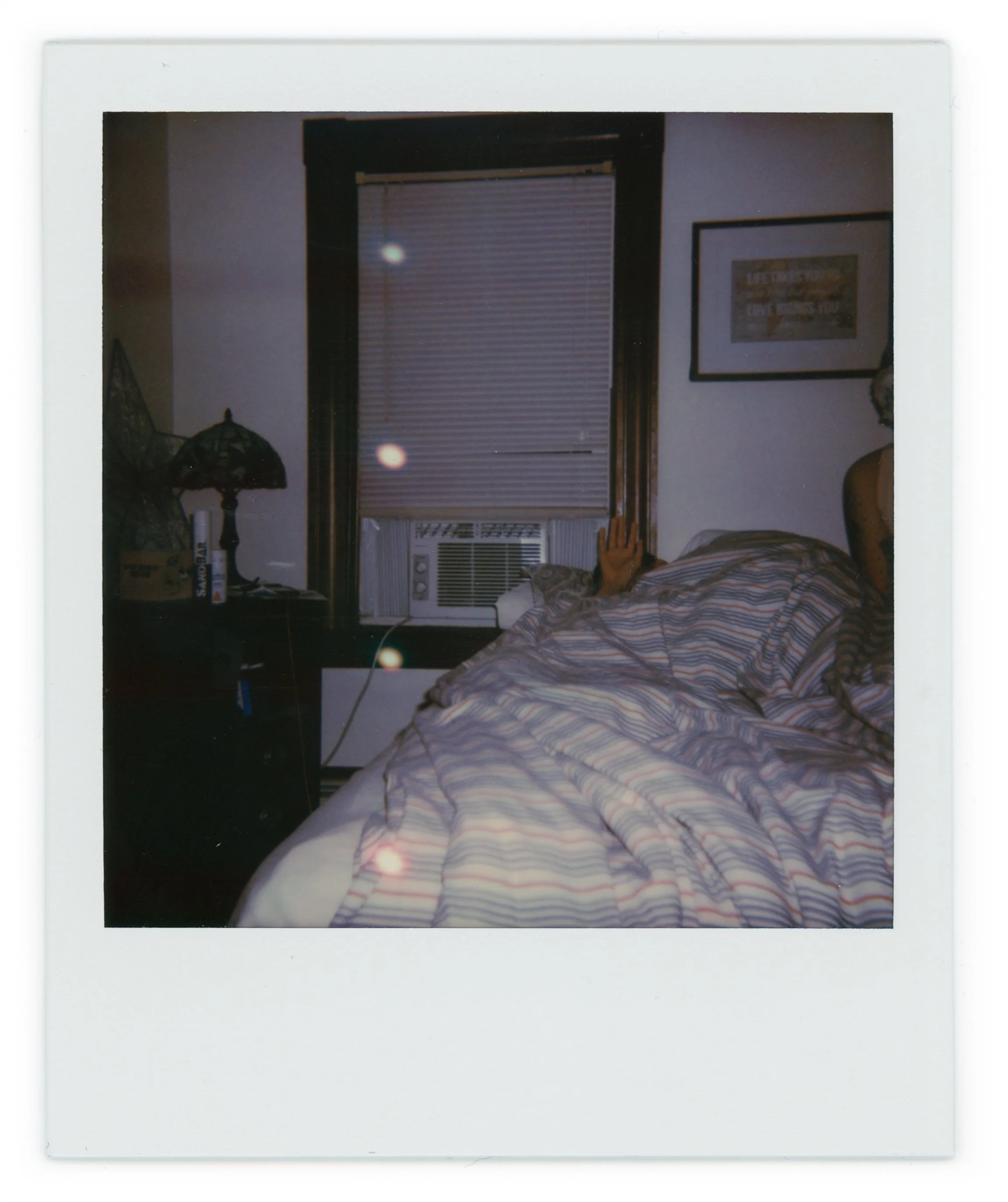 A person lying in bed wearing a striped shirt, with an arm reaching out from under the covers, in a dimly lit room with a window air conditioning unit and a framed picture on the wall.