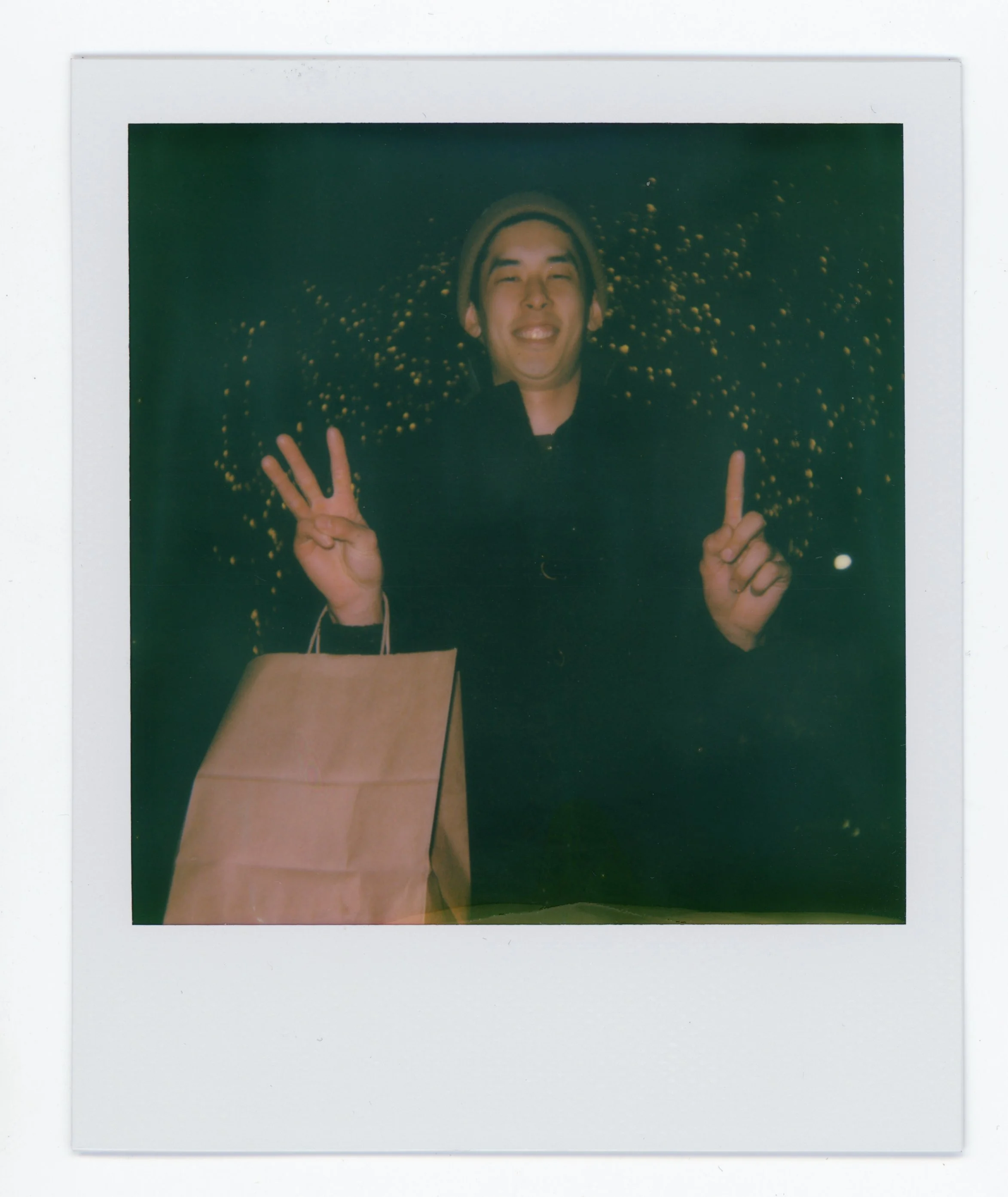 Person smiling and making peace signs with both hands, holding a shopping bag, in front of a decorated Christmas tree at night.