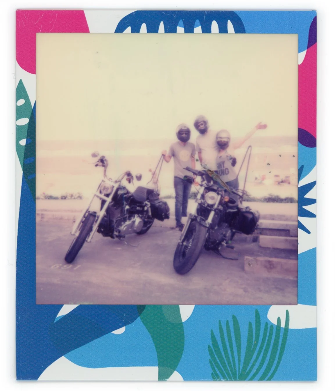 A vintage photo of three people standing outdoors next to two motorcycles, with one person raising an arm, on a boardwalk near the beach.