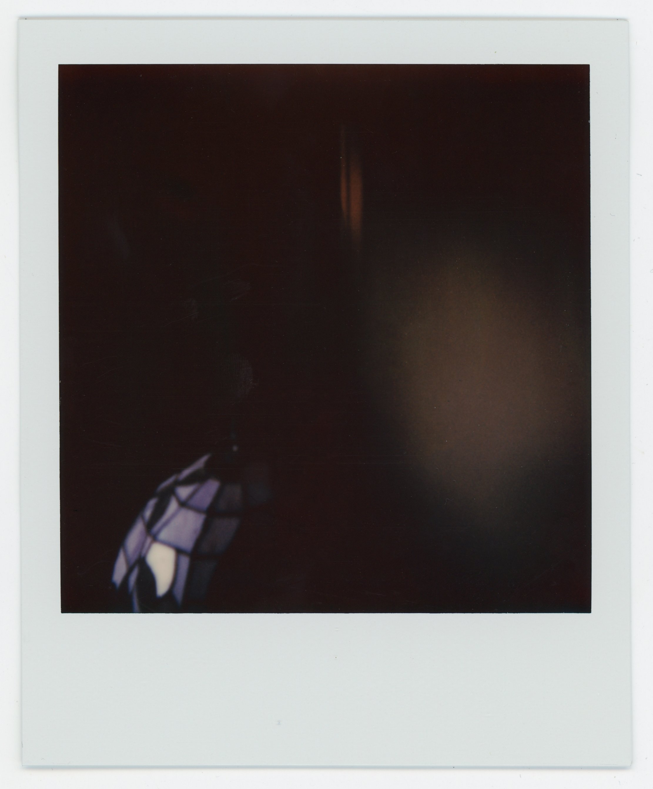 A dark Polaroid photo with a faint light on the top and a stained glass lamp in the bottom left corner.