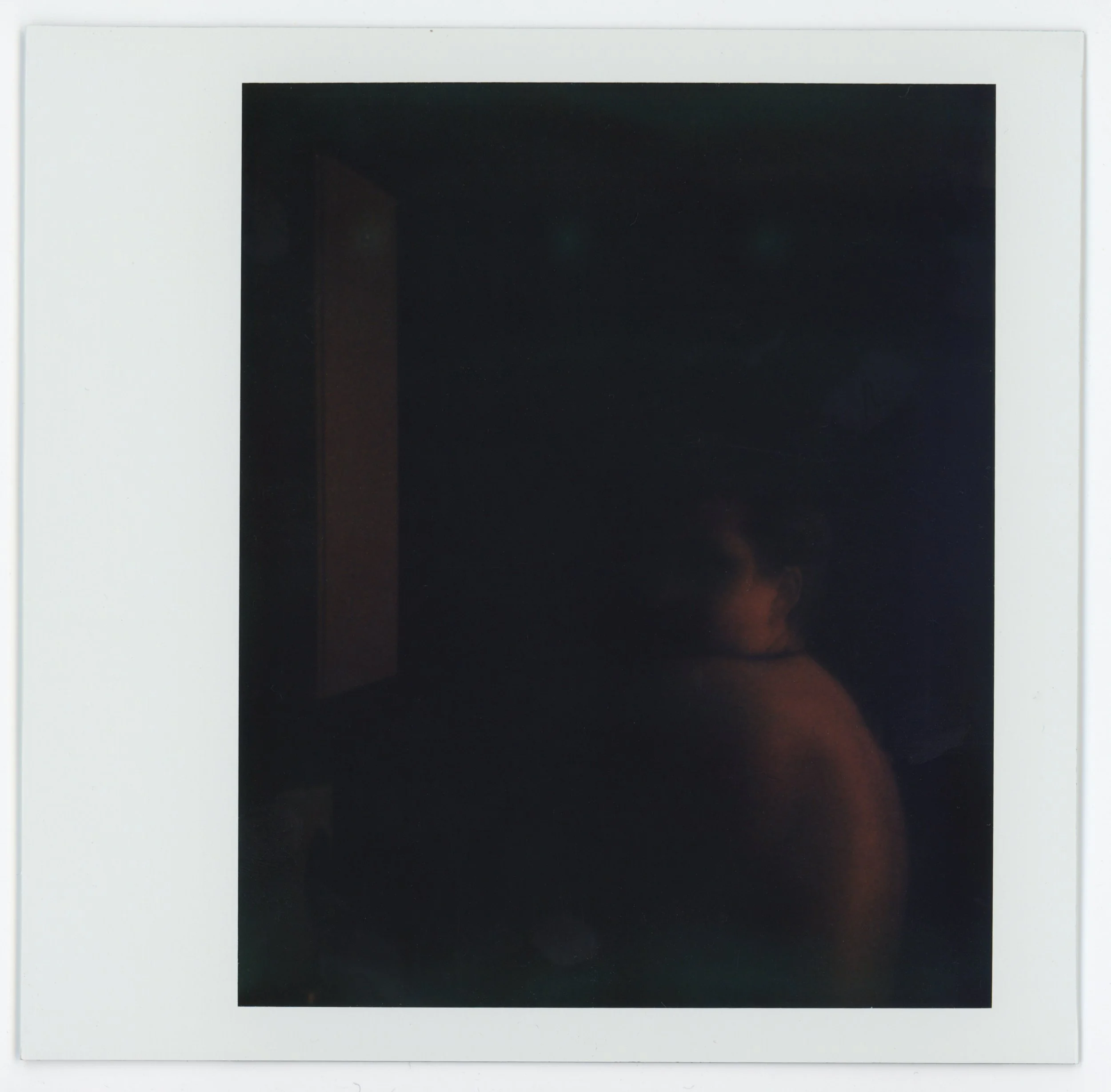 Dark Polaroid photograph of a person with glasses and short hair, looking to the side, with their shoulder visible, in a dimly lit environment.