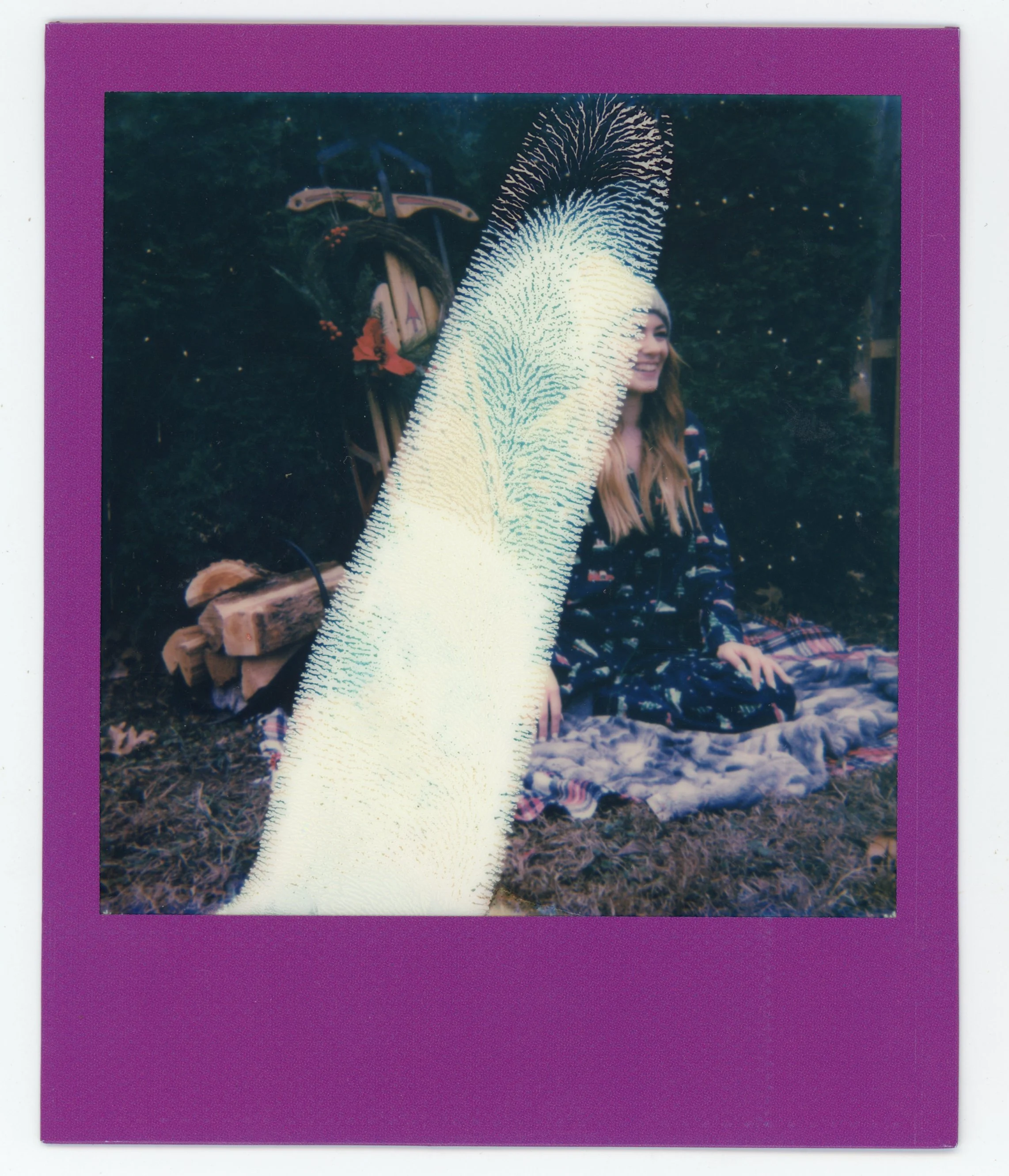 A woman sitting on a blanket outdoors with tall grass and trees in the background, smiling, with a bicycle decorated with flowers behind her. A large, light-colored furry animal or object partially covering the photo is in the foreground.