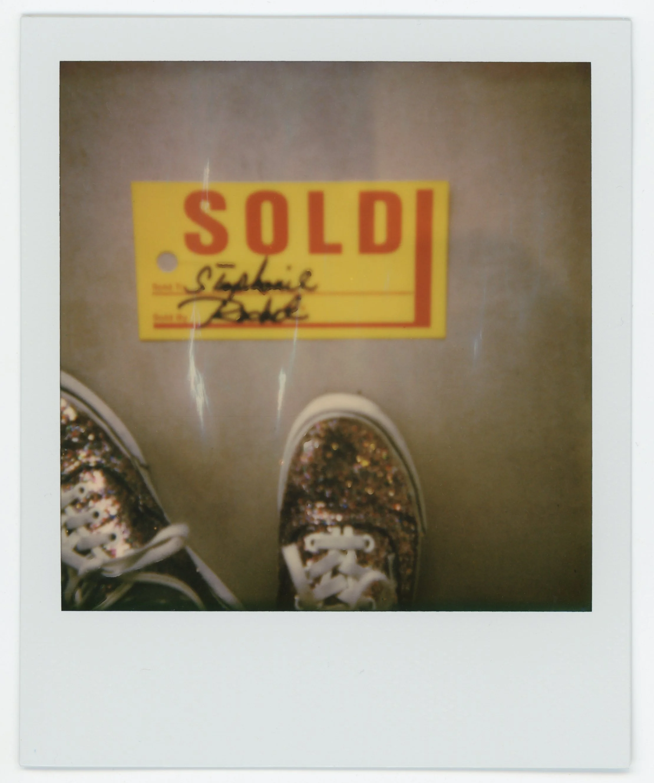 Polaroid photo showing a pair of glittery shoes on a gray floor next to a yellow sign with red text that reads 'SOLD,' and handwritten text 'Stephanie' and an illegible signature.