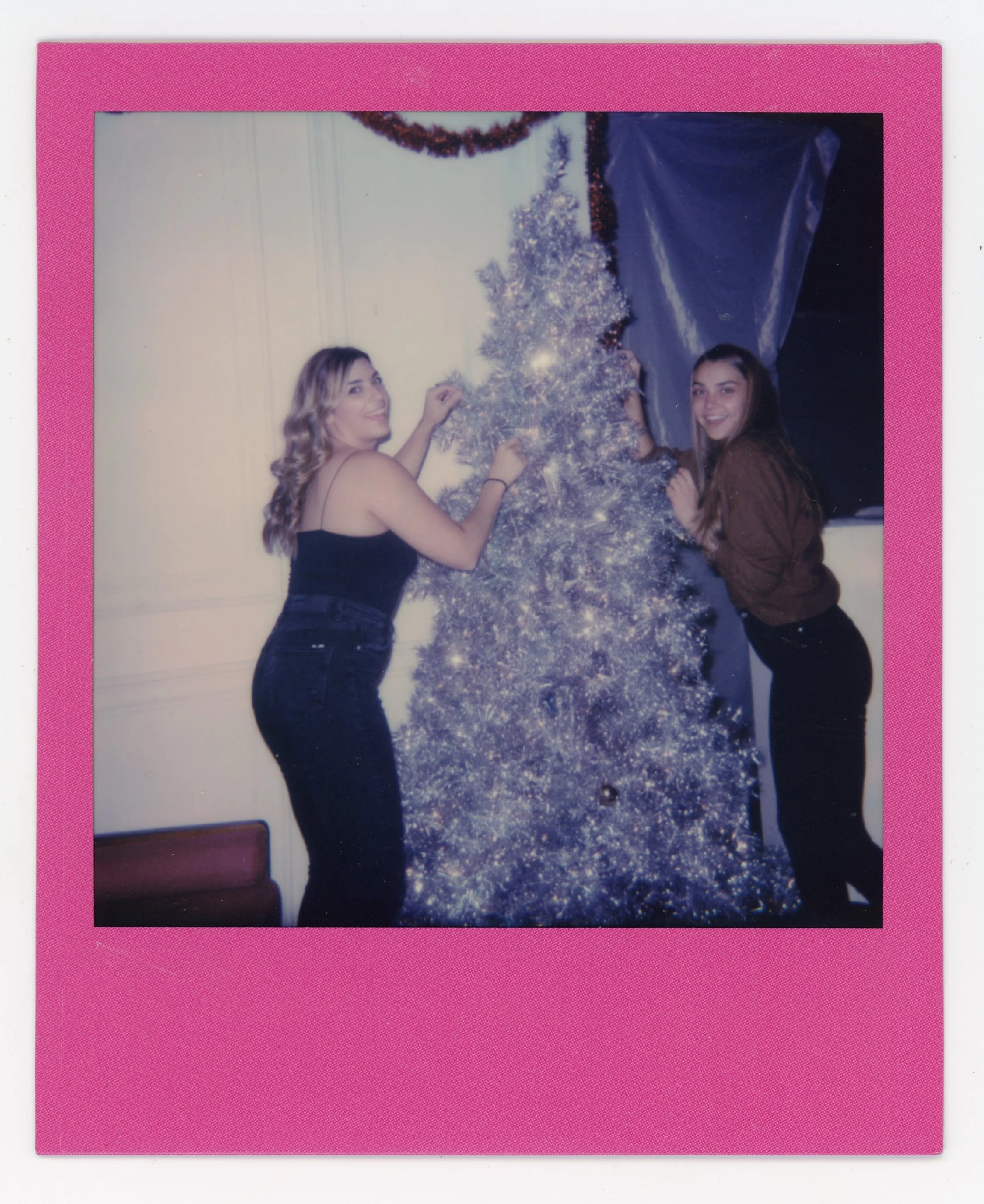 Two women decorating a Christmas tree with ornaments, standing on either side of the tree, inside a room with white walls and partially visible window with blue curtain, all framed with a pink border.