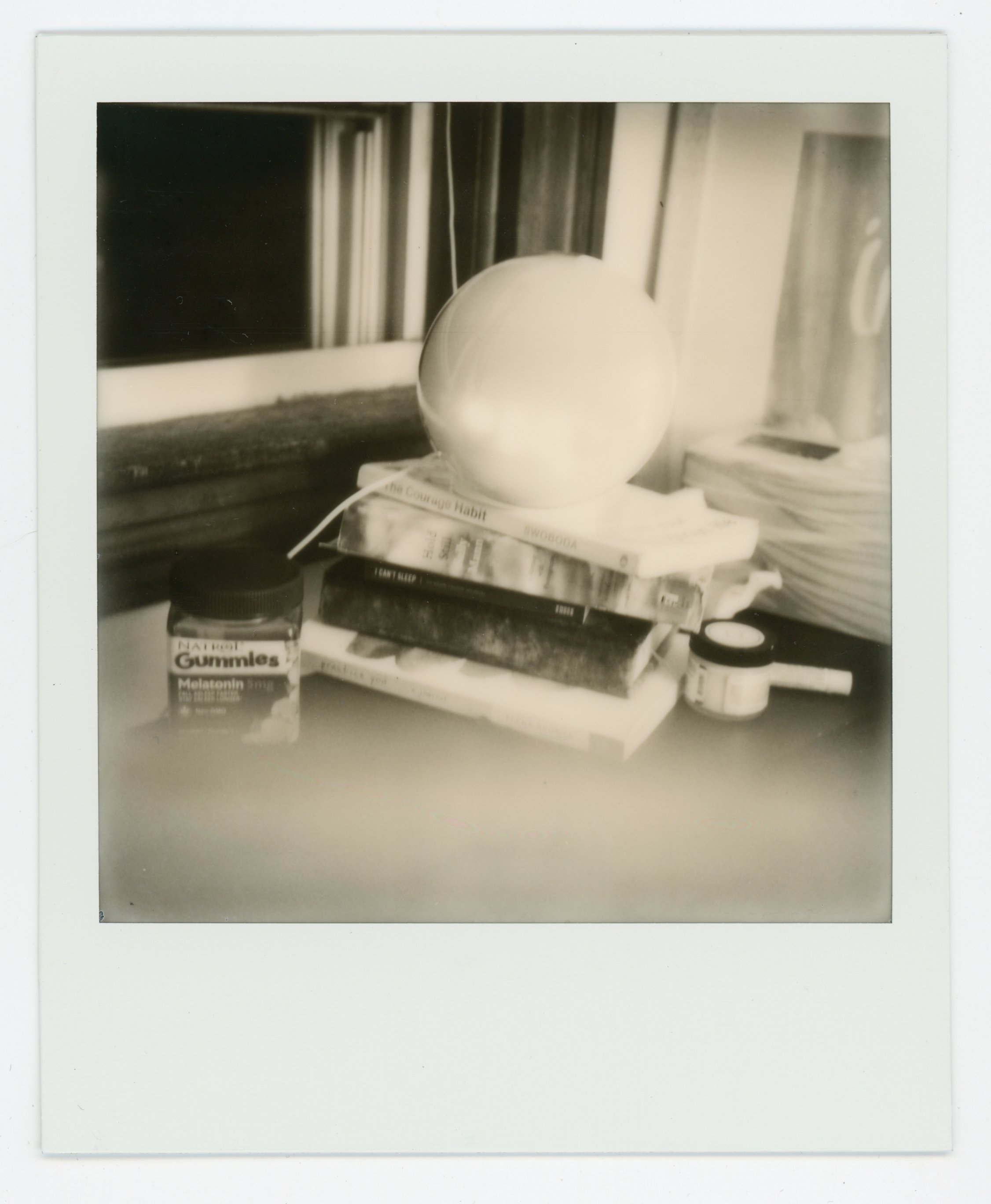 Black and white photo of a desk with books, a large crystal ball, a container of melatonin gummies, a small jar, a deck of cards, and a tub of cream, with a window in the background.