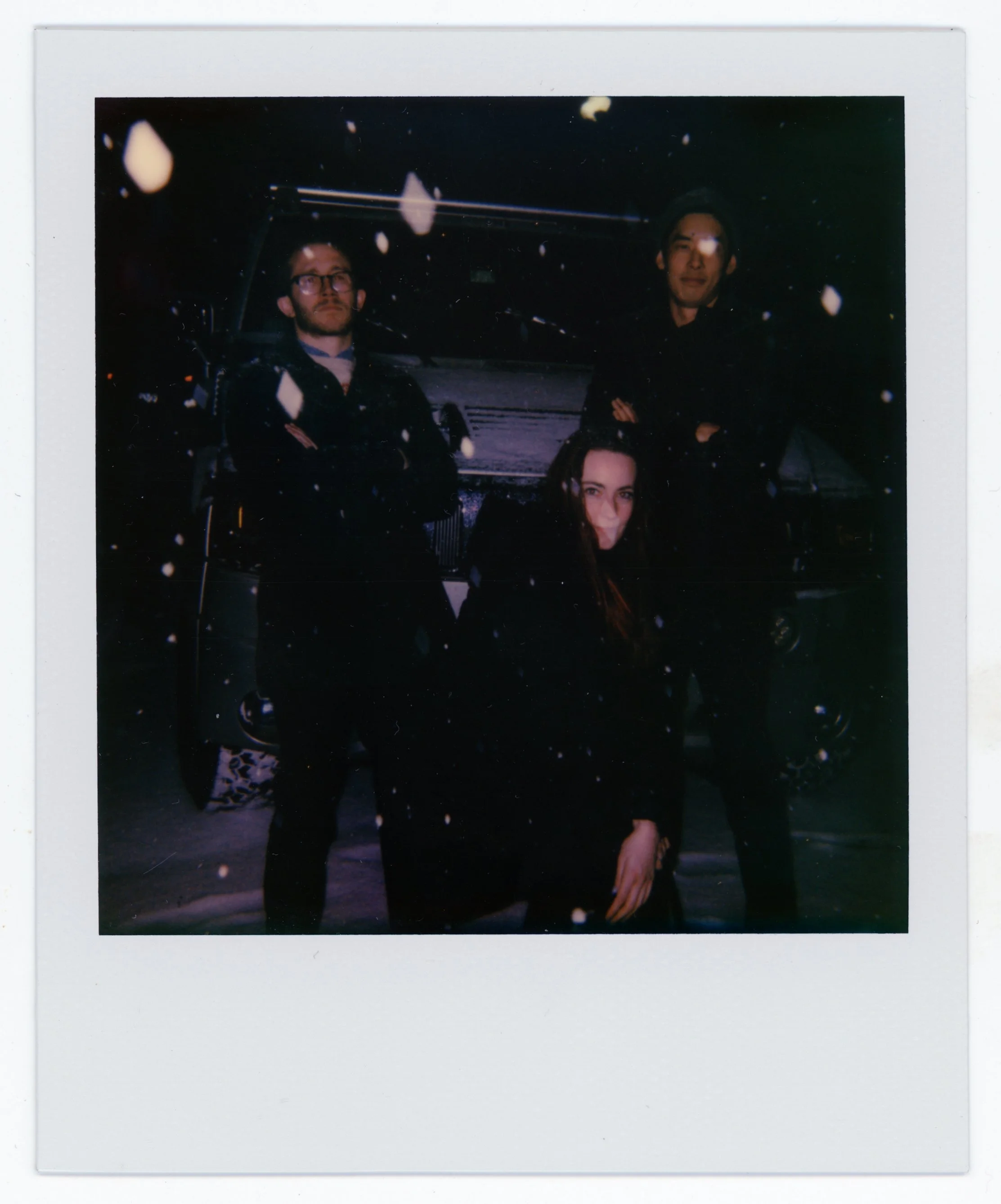 Three people posing outdoors at night with snow falling, standing in front of a vehicle.