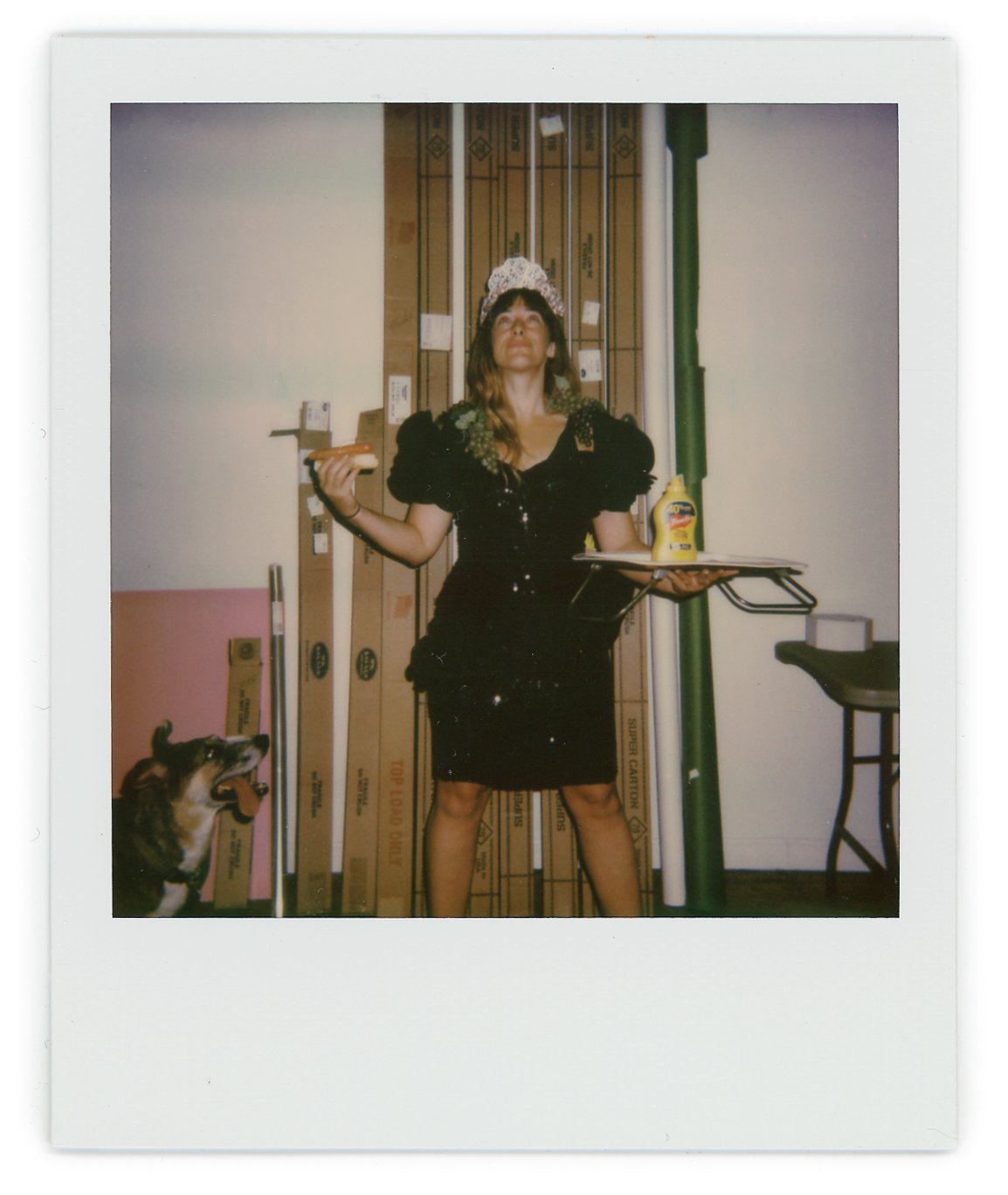 A woman dressed as a firefighter, wearing a tiara and a black dress, holding a hot dog in one hand and a tray with mustard in the other, standing indoors with a dog looking up at her.