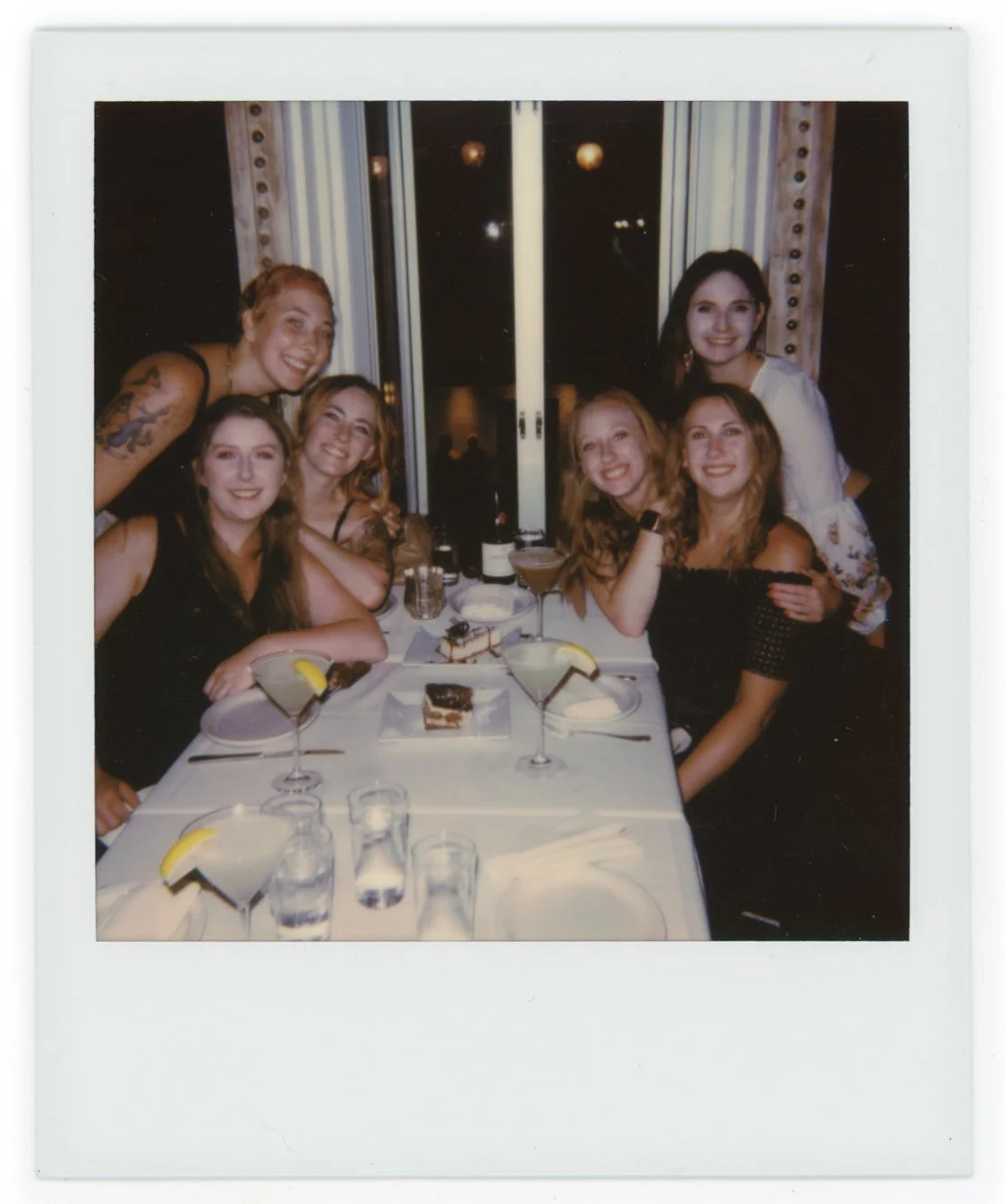Group of six women smiling around a dinner table with plates, desserts, and drinks, at night in a restaurant or event space.