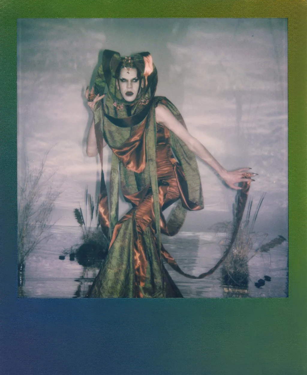 A person dressed in elaborate, flowing costume with earthy tones stands in front of a painted background resembling a marsh or wetland, with tall grasses and water, and makes an expressive gesture.