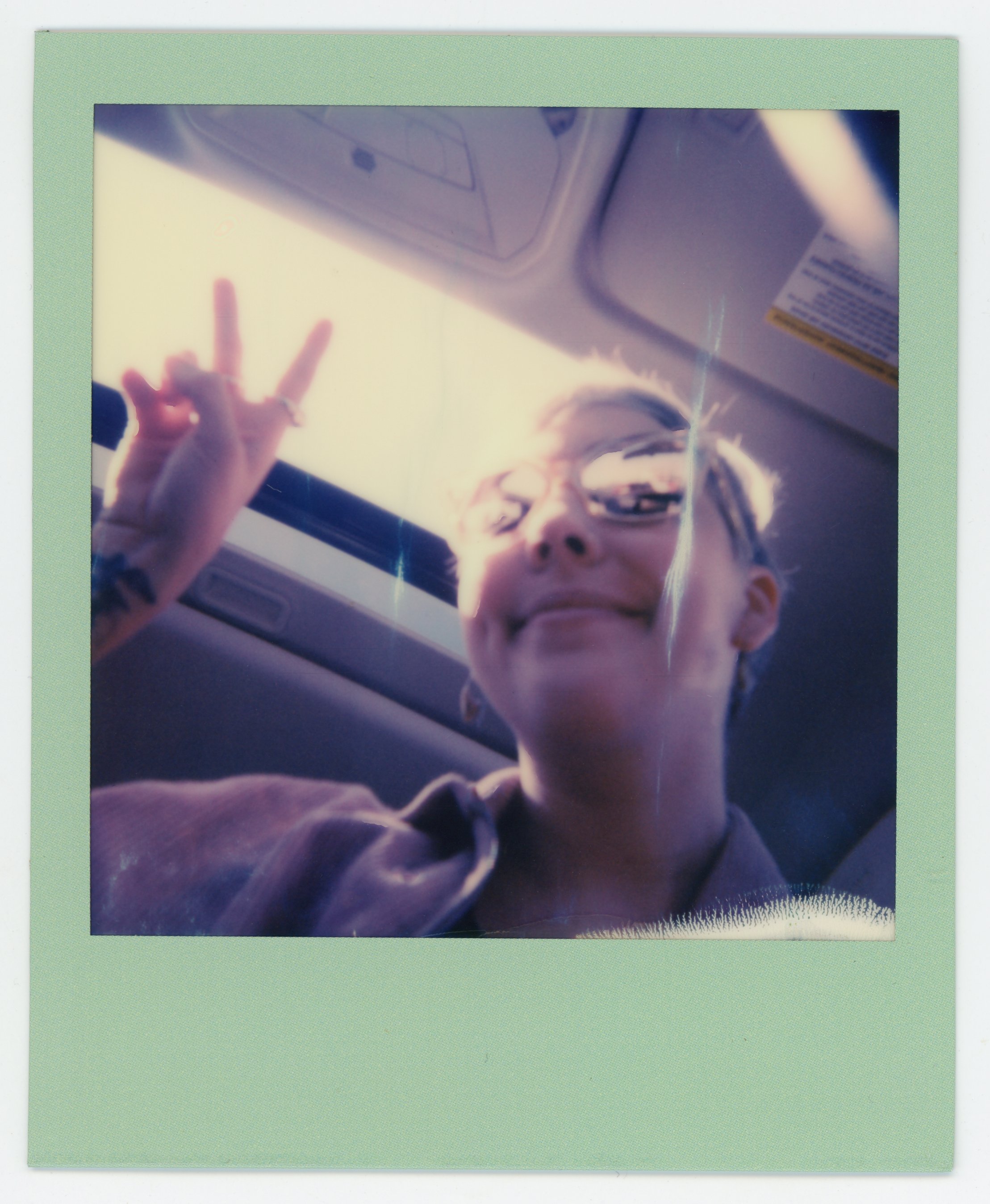 A person with glasses and a braid smiling and making a peace sign while sitting inside a vehicle, with sunlight streaming through the window.