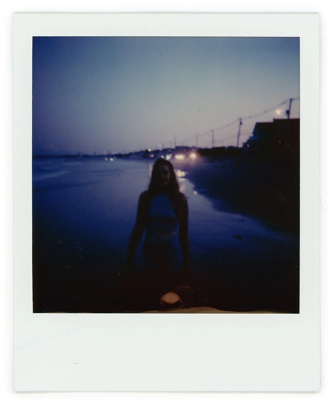 A blurry Polaroid photo of a woman standing on a beach at dusk with the ocean behind her and lights in the distance.