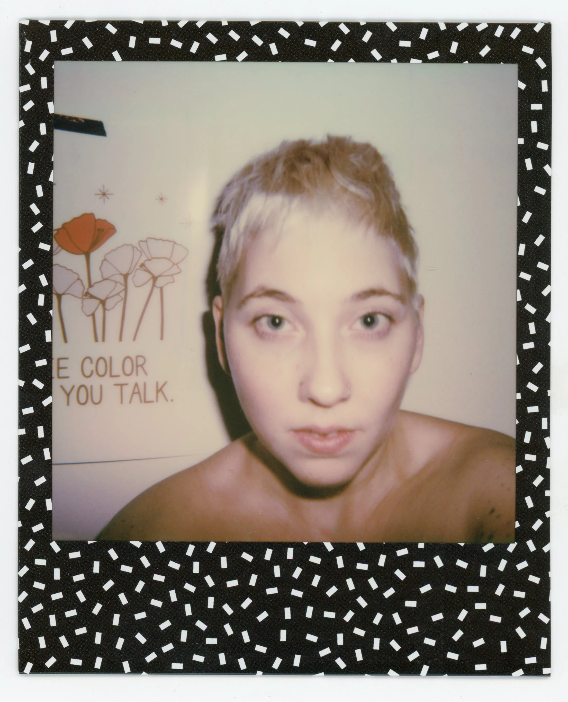 A young woman with short, blonde hair and light skin takes a Polaroid selfie, with a white wall in the background featuring artwork of flowers and the text 'THE COLOR YOU TALK.'