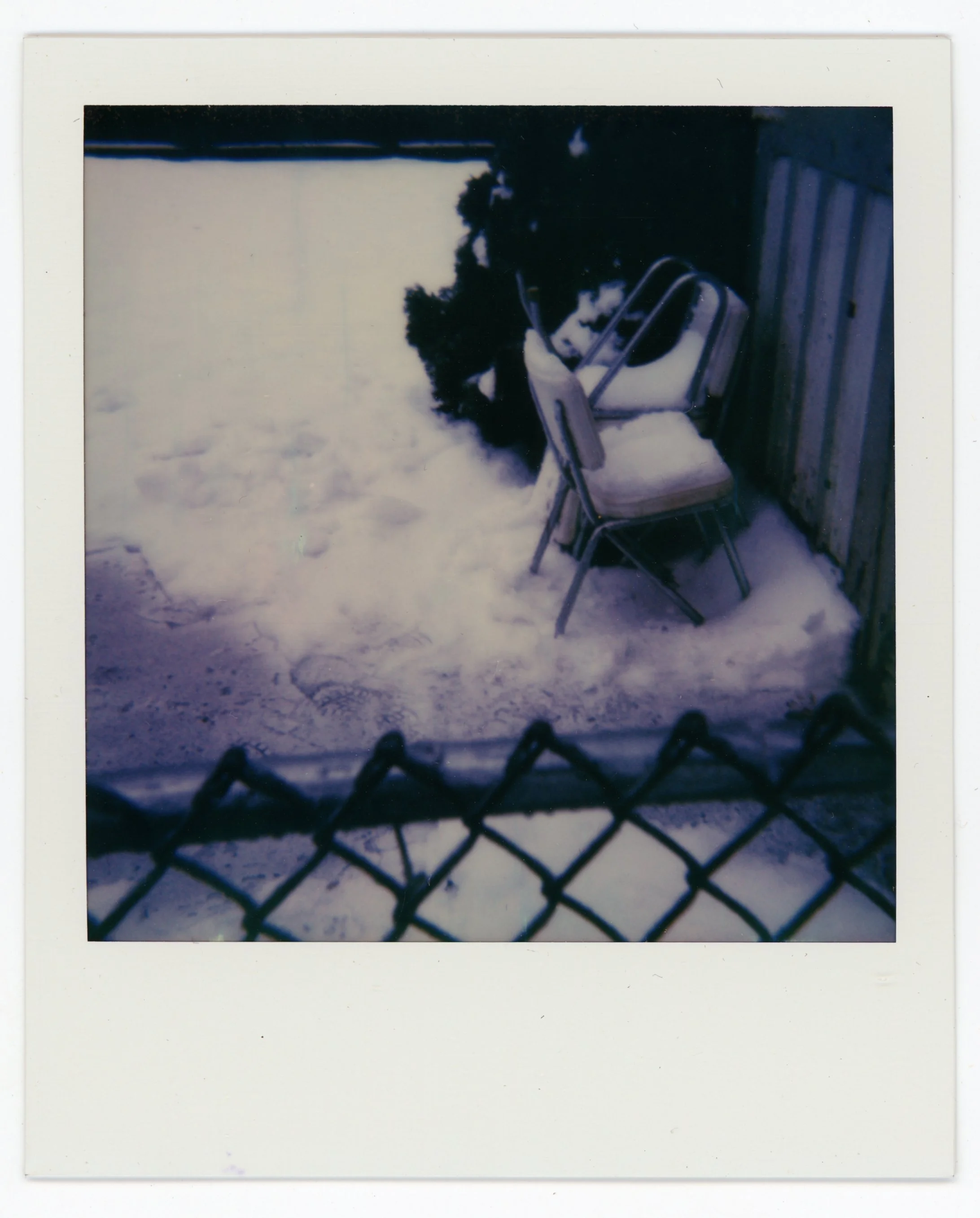 Snow-covered outdoor area with a chain-link fence at the foreground, leaning chairs around a stone wall, and snow-covered ground.