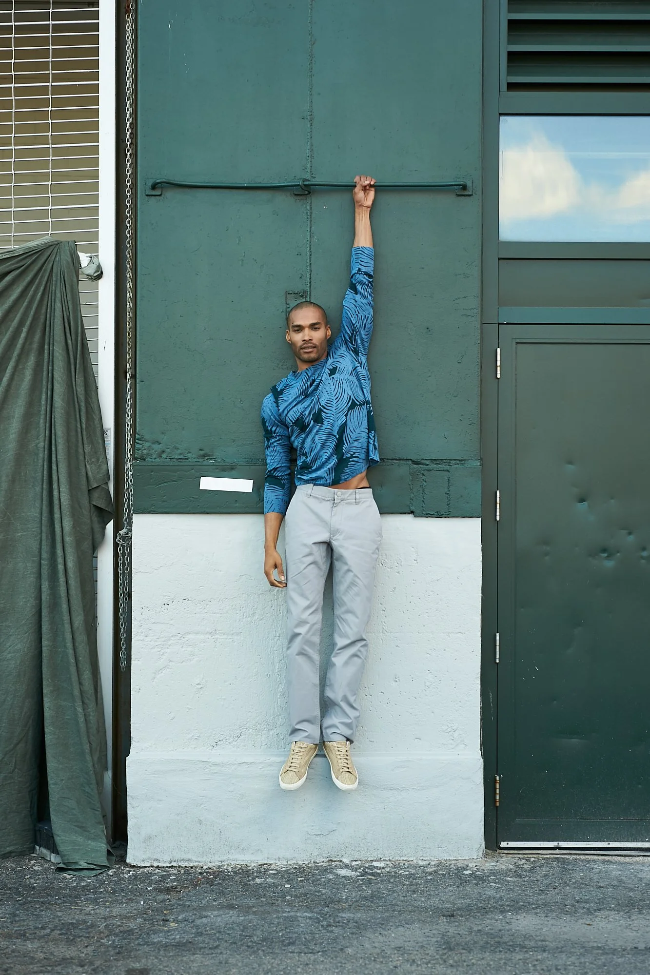 A man hanging from a horizontal bar on a green wall outside, with his feet just off the ground, wearing a blue patterned shirt, light gray pants, and beige sneakers.