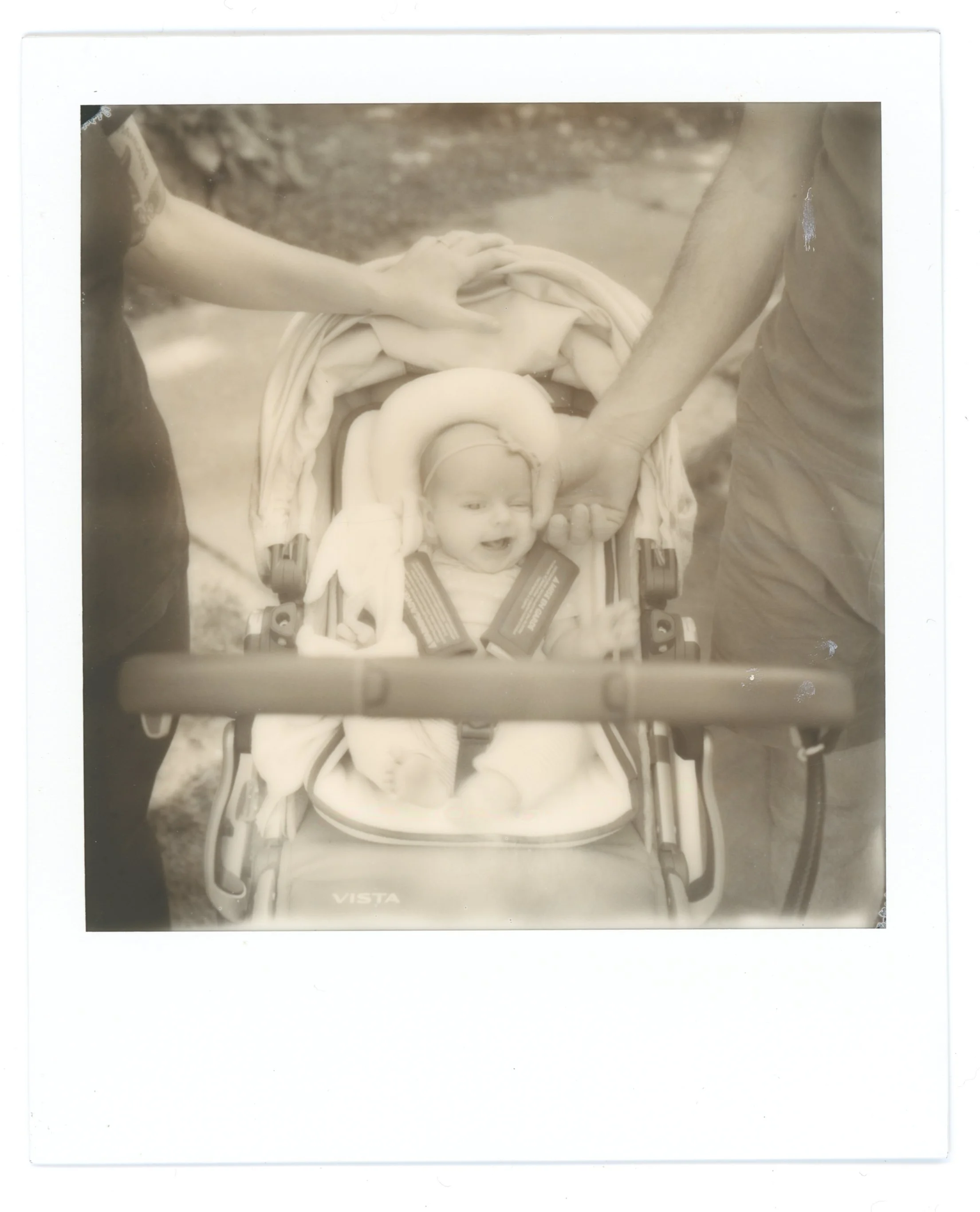 Black and white photo of a smiling baby in a stroller, held by two people whose hands are visible, outdoors.