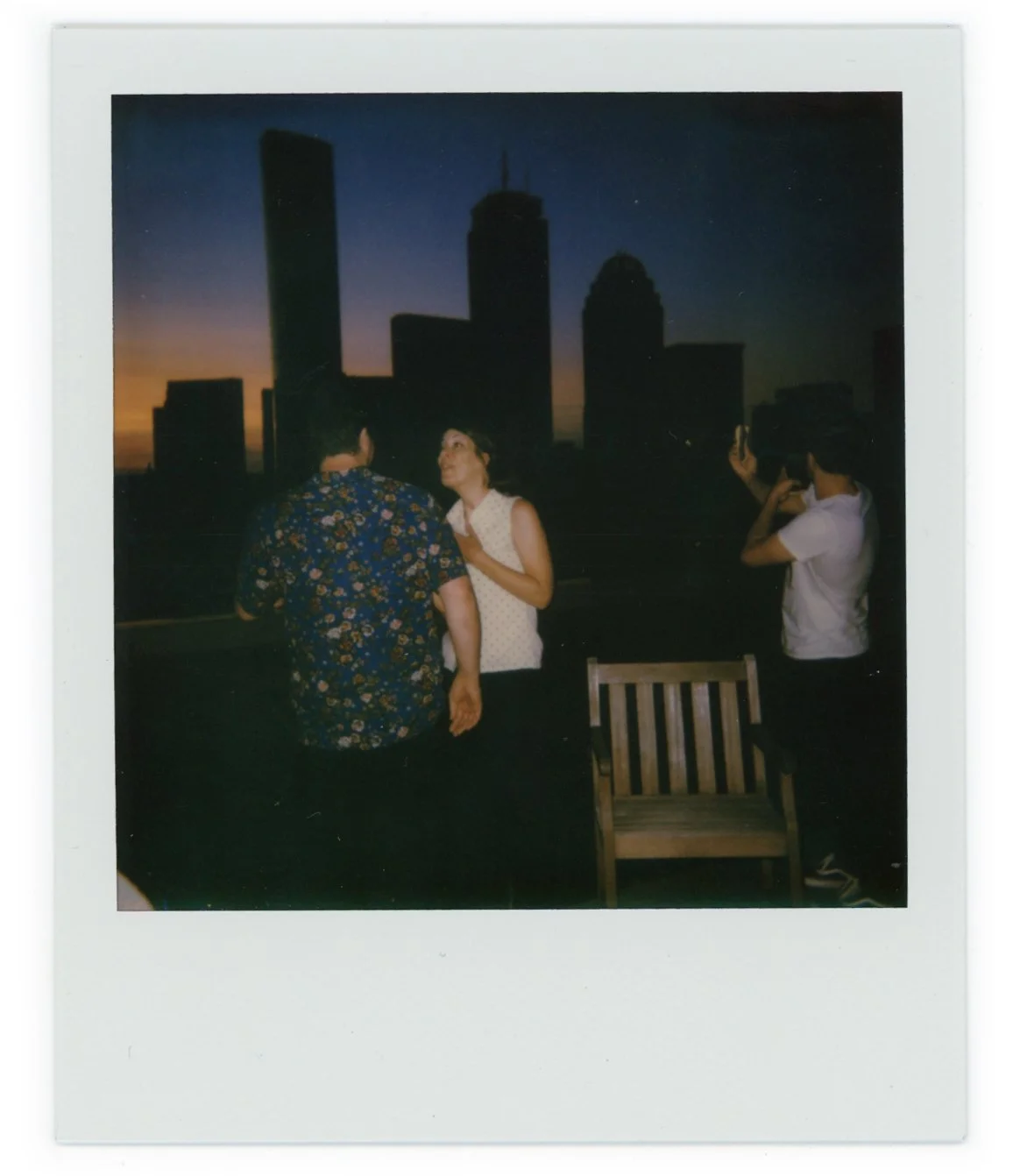 Three people on a rooftop during sunset, with city skyscrapers in the background. Two are talking, and one is taking a photo.