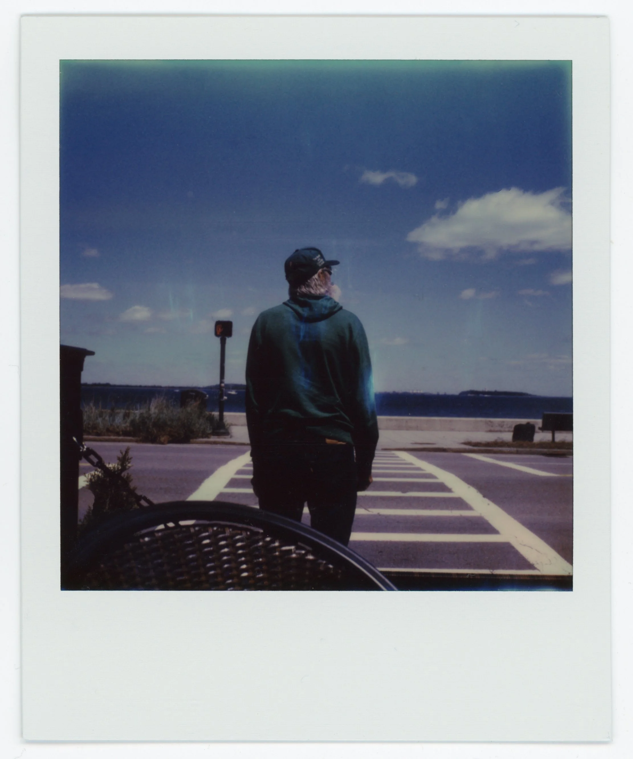 A person with a hoodie and cap standing on a crosswalk near a body of water, with a partly cloudy sky overhead.