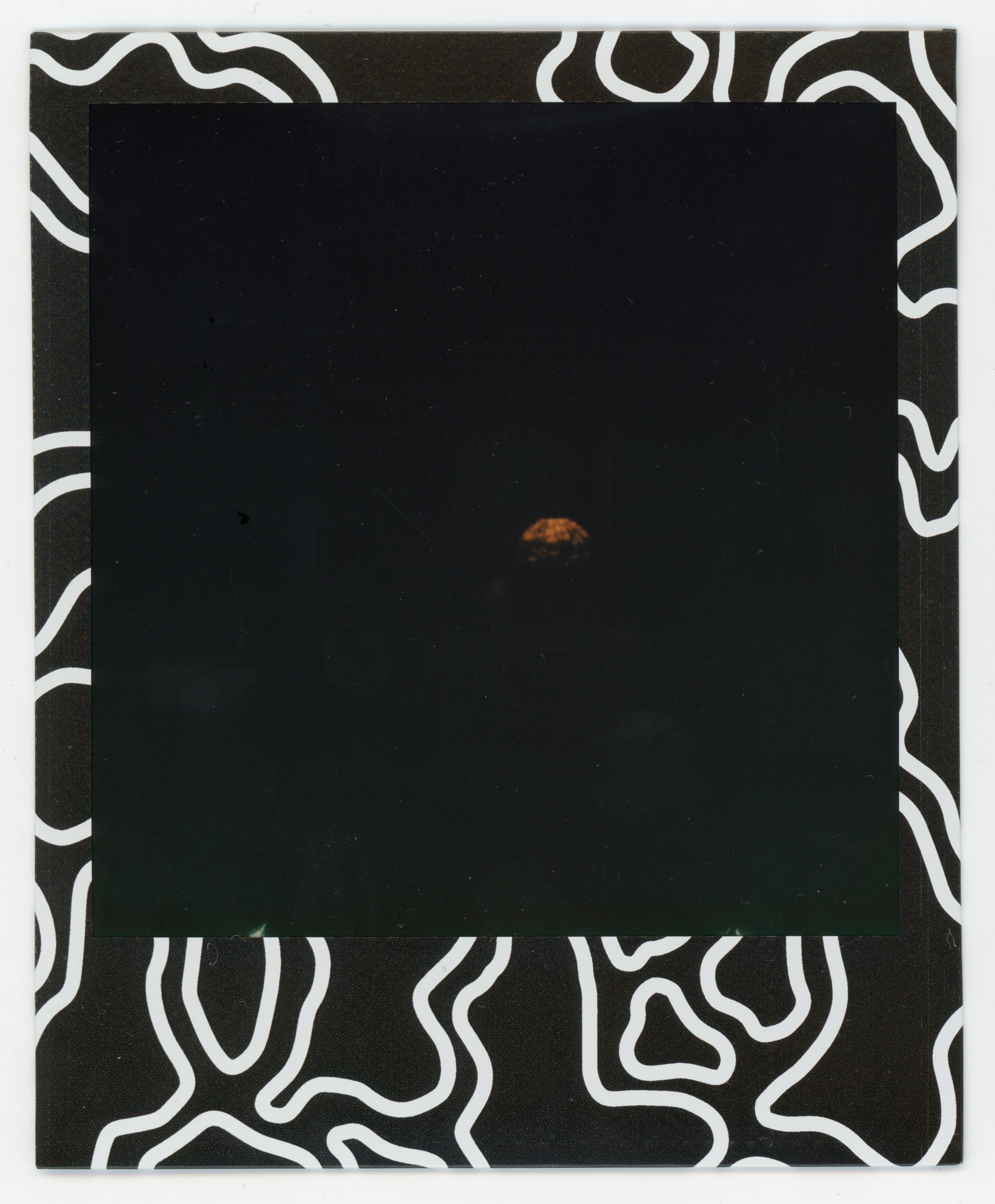 The image is a Polaroid photo with an abstract black and white patterned border. Inside the Polaroid, there's a very dark scene with a small, faintly illuminated object or structure in the center, barely visible against the dark background.