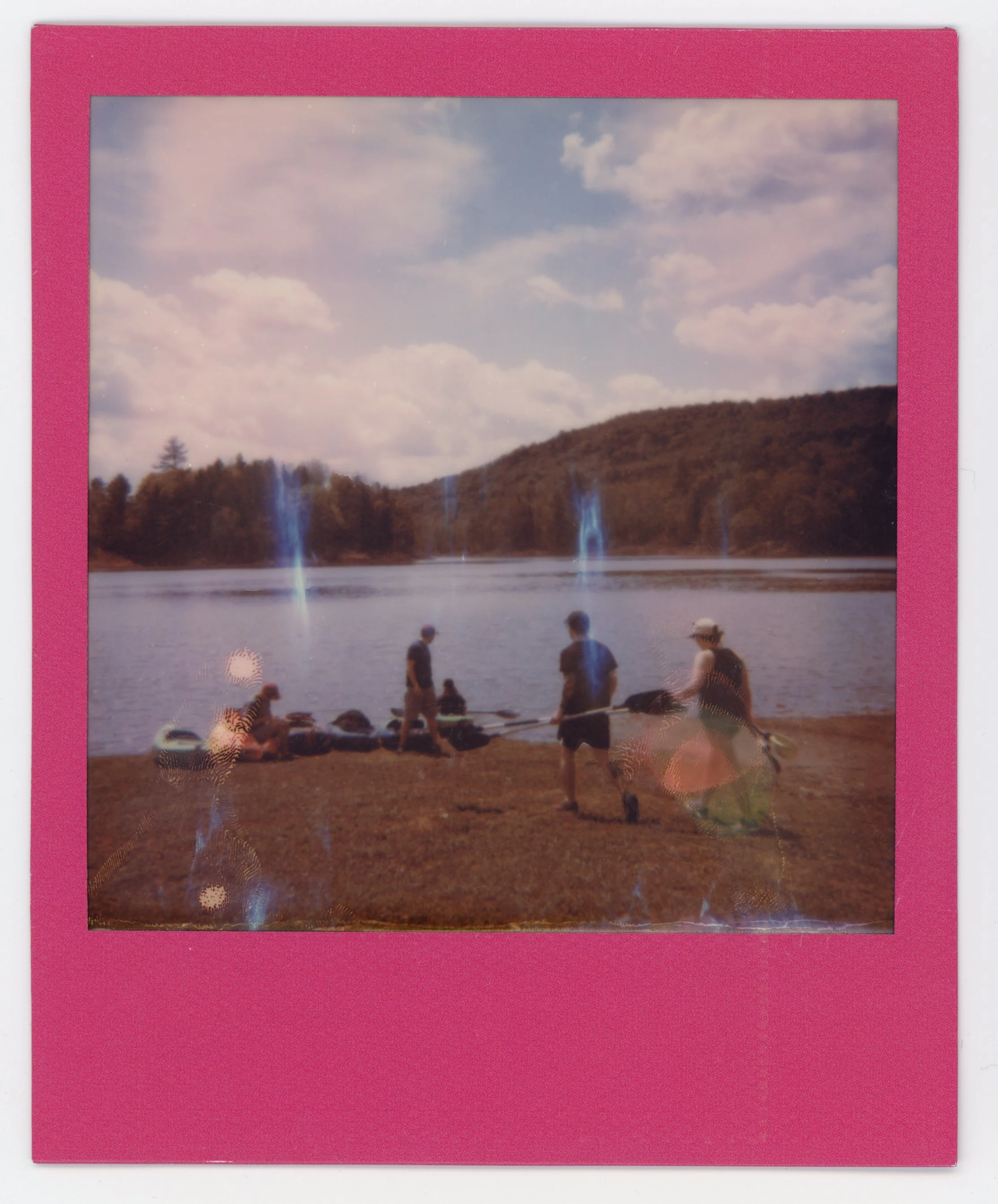 People preparing to go kayaking by a lake with blue waterfalls in the background, trees, grassy shore, and partly cloudy sky.