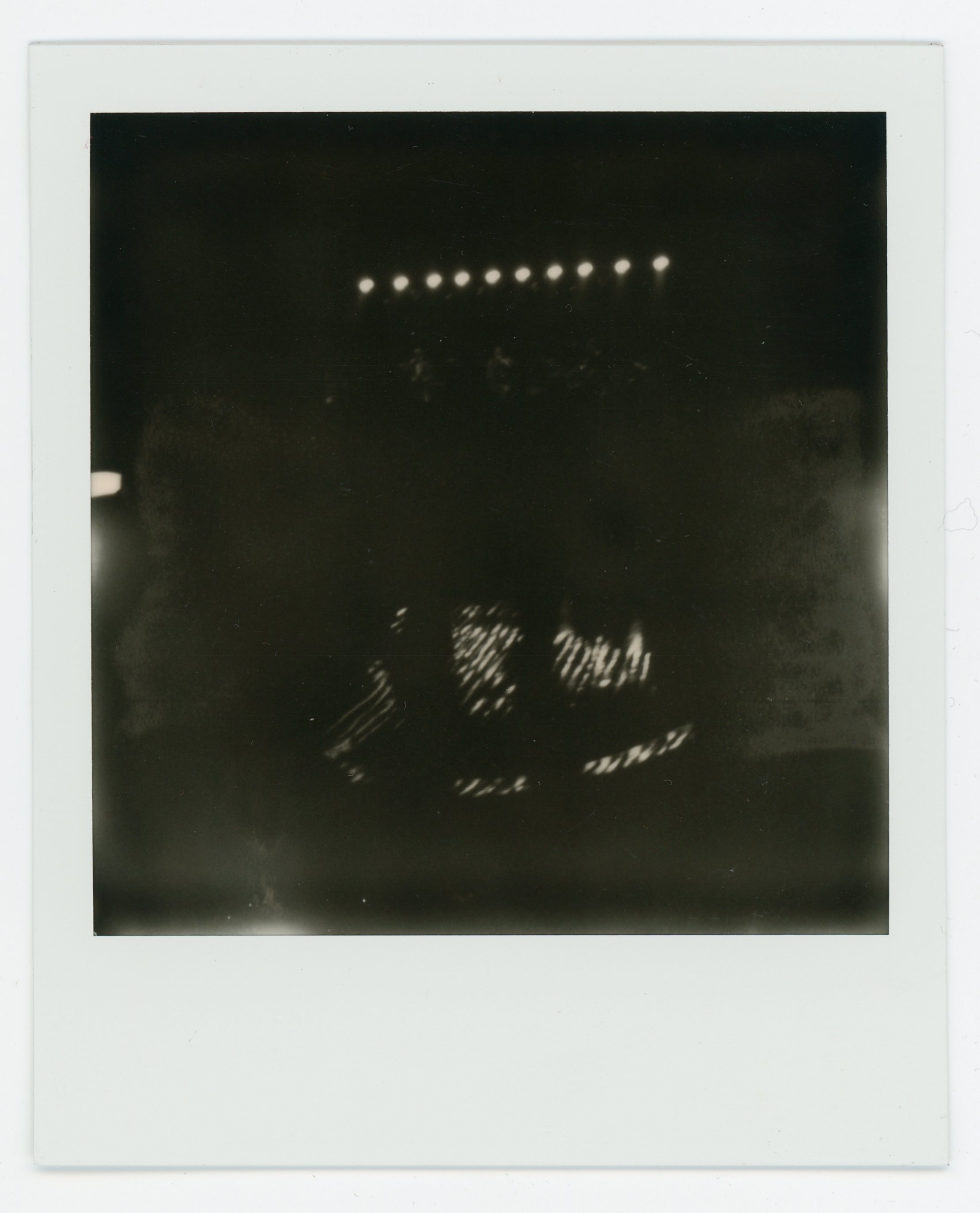 Black and white Polaroid photo of a dark scene with illuminated stage lights and blurred figures or objects.