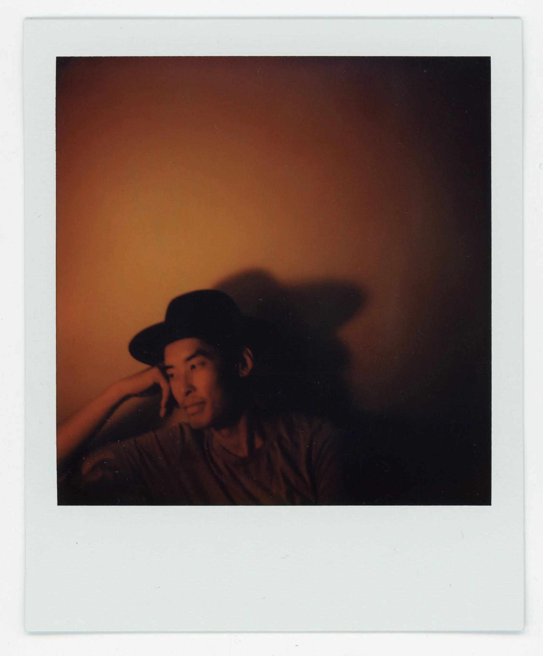 A man wearing a wide-brimmed hat and a striped shirt, smiling and resting his head on his hand, in a dimly lit setting with an orange background and shadow on the wall.