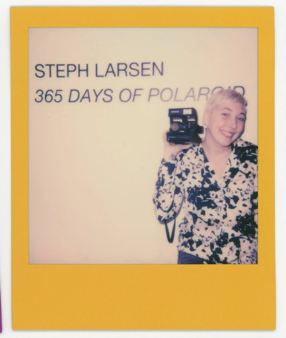 A woman with short blonde hair smiling and holding a vintage camera, standing in front of a sign that reads "Steph Larsen 365 Days of Polaroid".