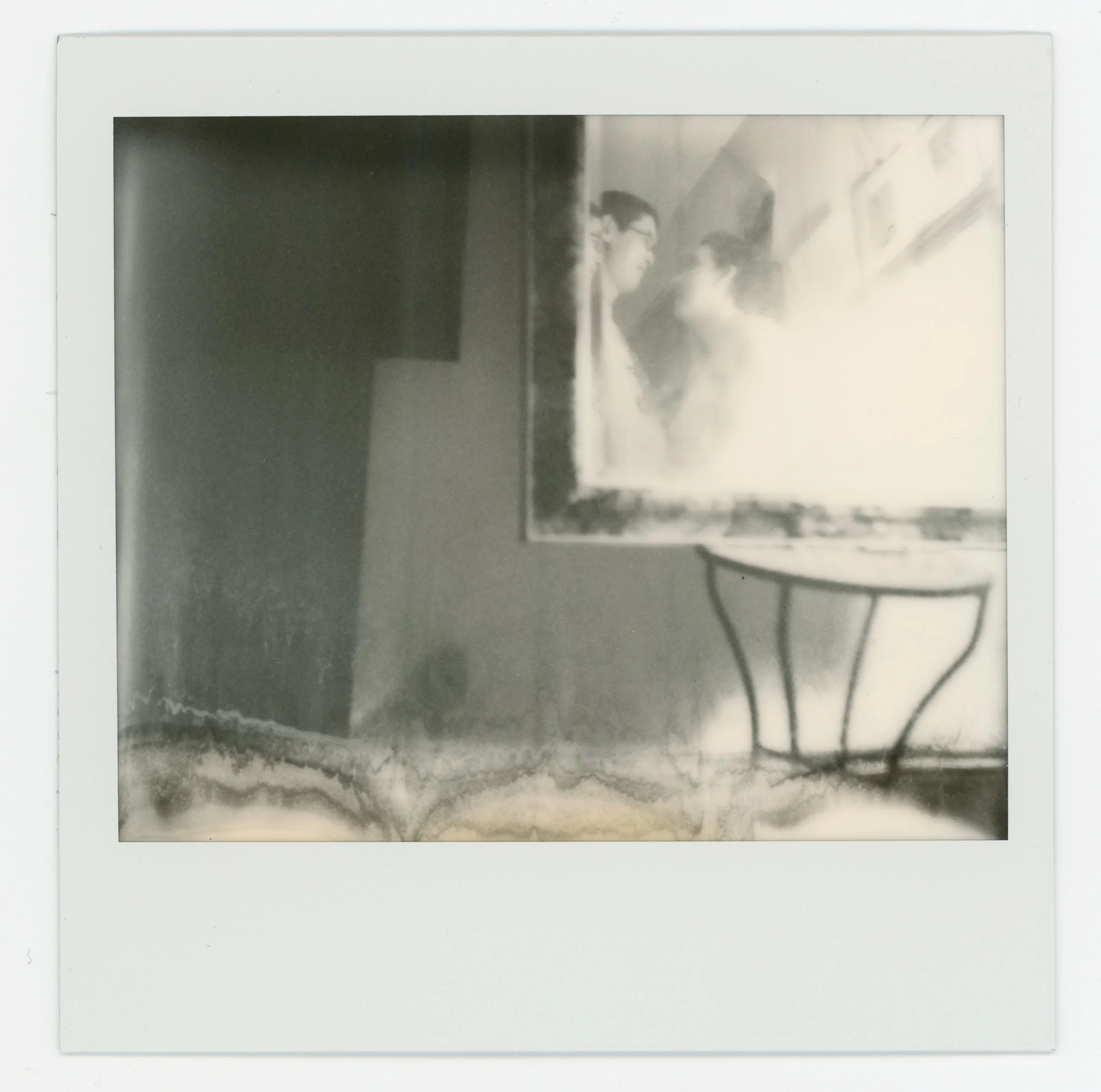 A black and white Polaroid photo featuring a reflective surface or glass on the right and a small table with curved legs below it.