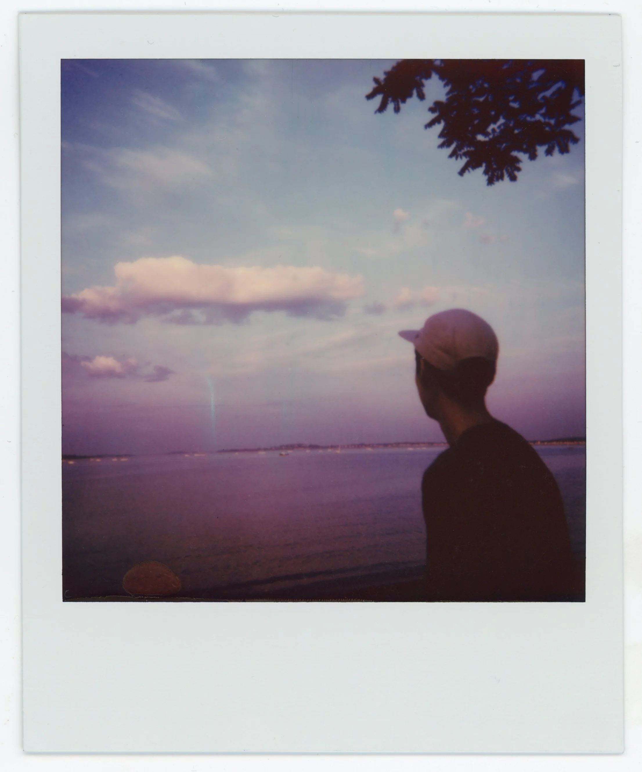 Person with a cap looking at sunset over a body of water, with clouds and a tree branch in the sky