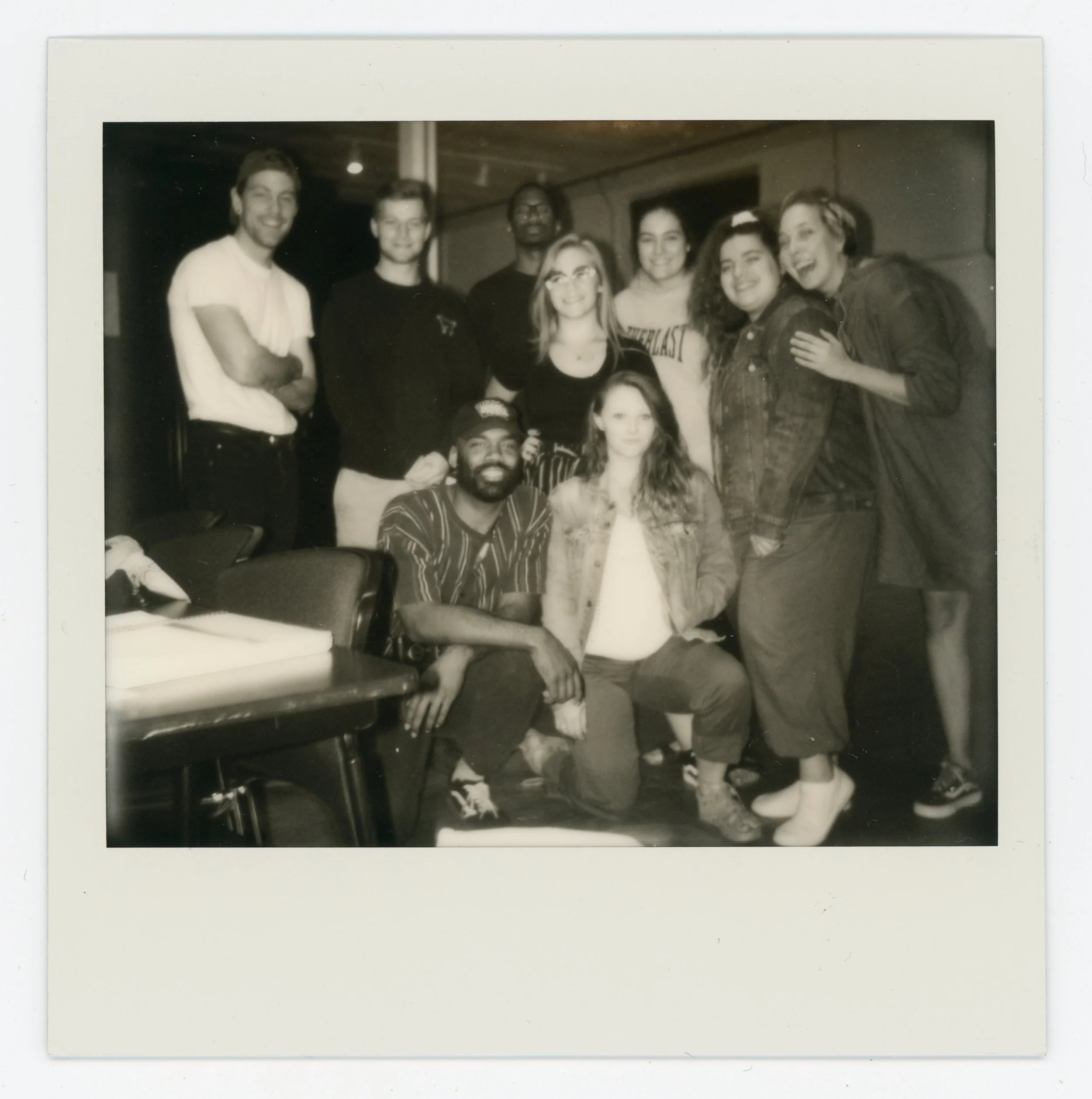 A group of nine diverse young adults posing together in black and white, some standing and some sitting, in an indoor setting with a casual atmosphere.