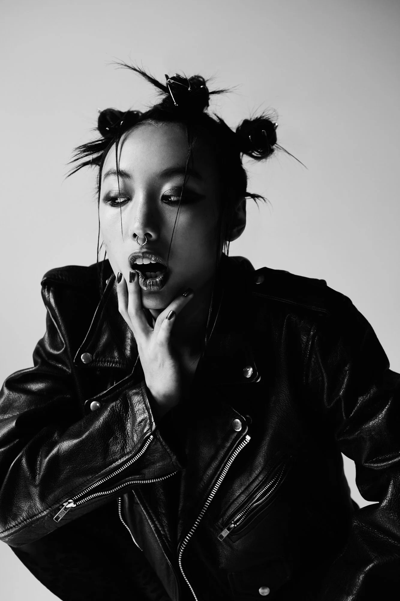 Black and white portrait of a woman with short, messy hair styled with small buns, wearing a black leather jacket, with her hand on her face, showing a surprised or intense expression.