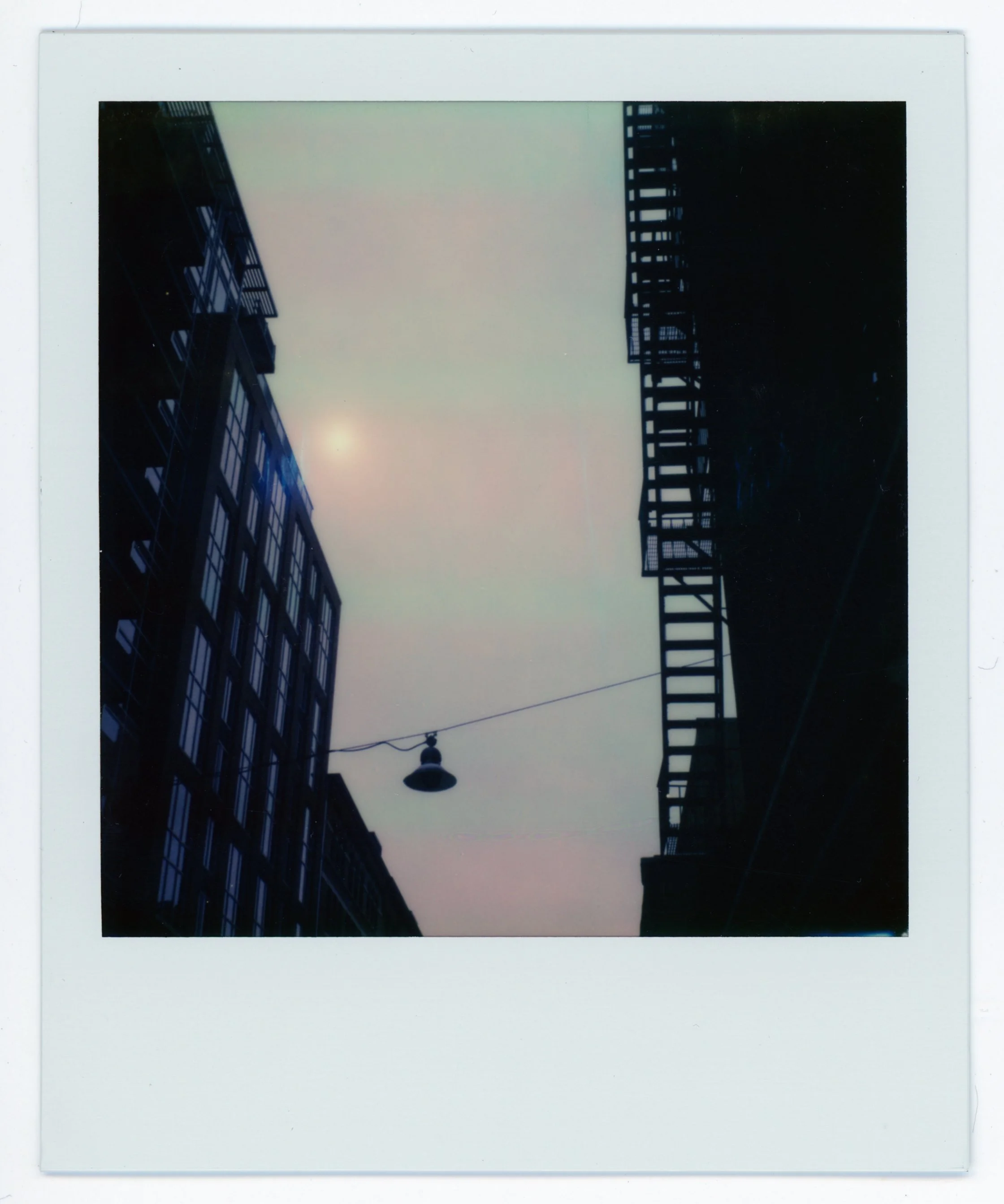 Silhouette of buildings with fire escape on a street, hanging lamp, and a hazy sun or moon in an overcast sky.