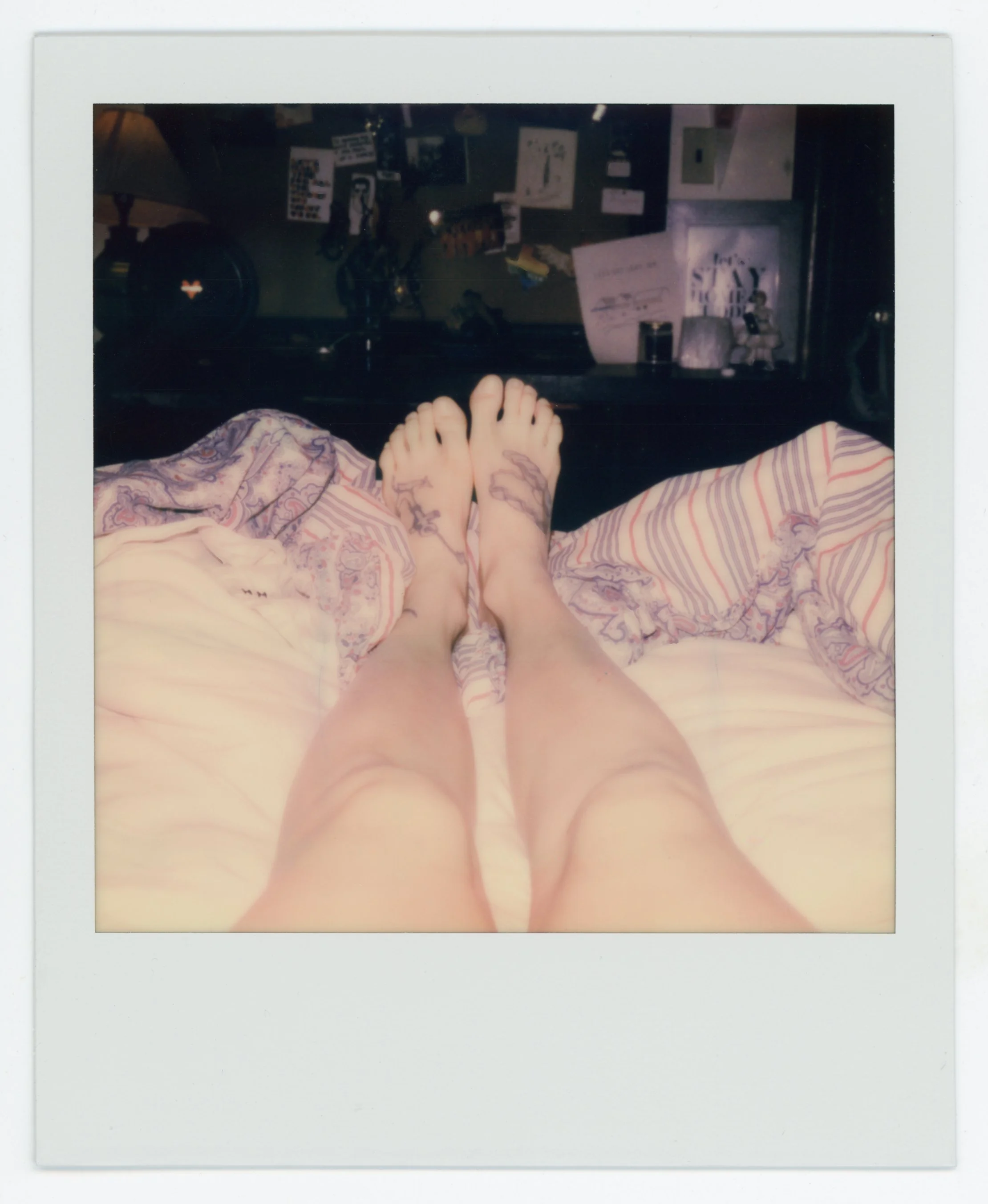A person with tattoos on both legs is lying in bed with legs slightly apart, taking a Polaroid-style photograph of their bare legs and feet. The bed has patterned sheets, and a nightstand with various items, including a lamp, is visible in the backgr