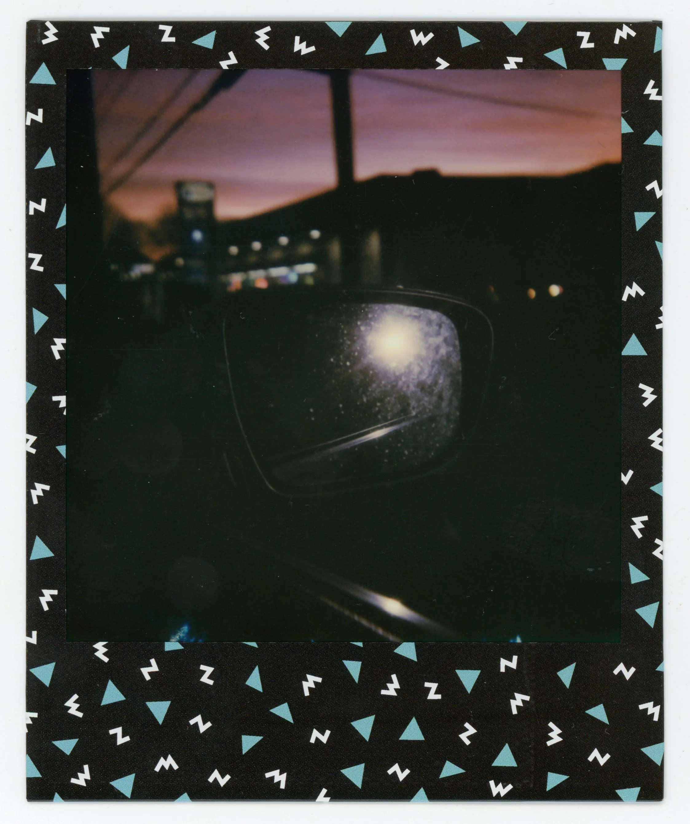 Photo of a car's side mirror reflecting a galaxy scene, with a sunset or dusk sky and city lights in the background, surrounded by a black frame with blue and white geometric shapes.