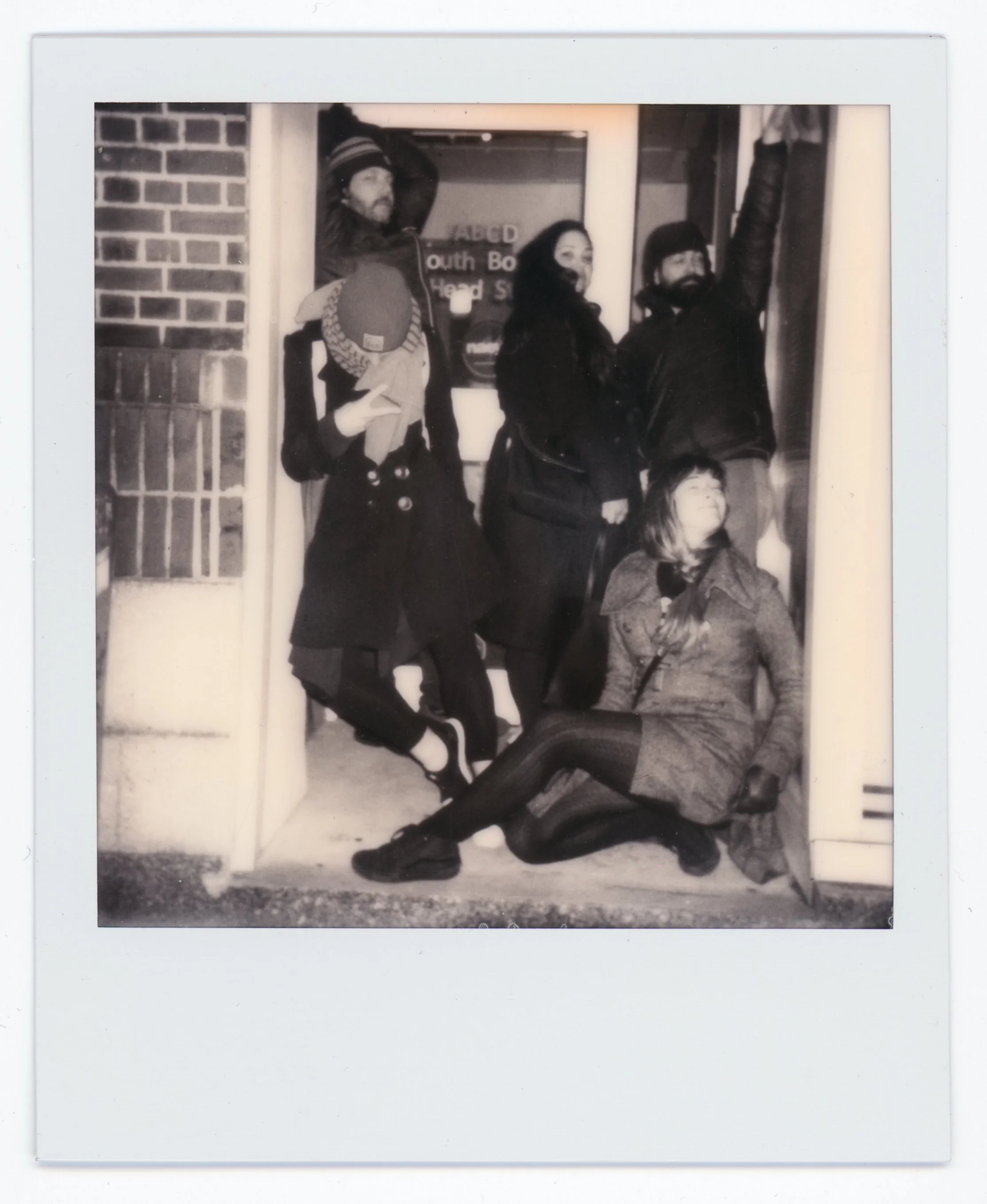 Group of five people posing on a doorstep outside a building, two standing, one seated on the ground, and two leaning against wall or window frame, in black and white photo.