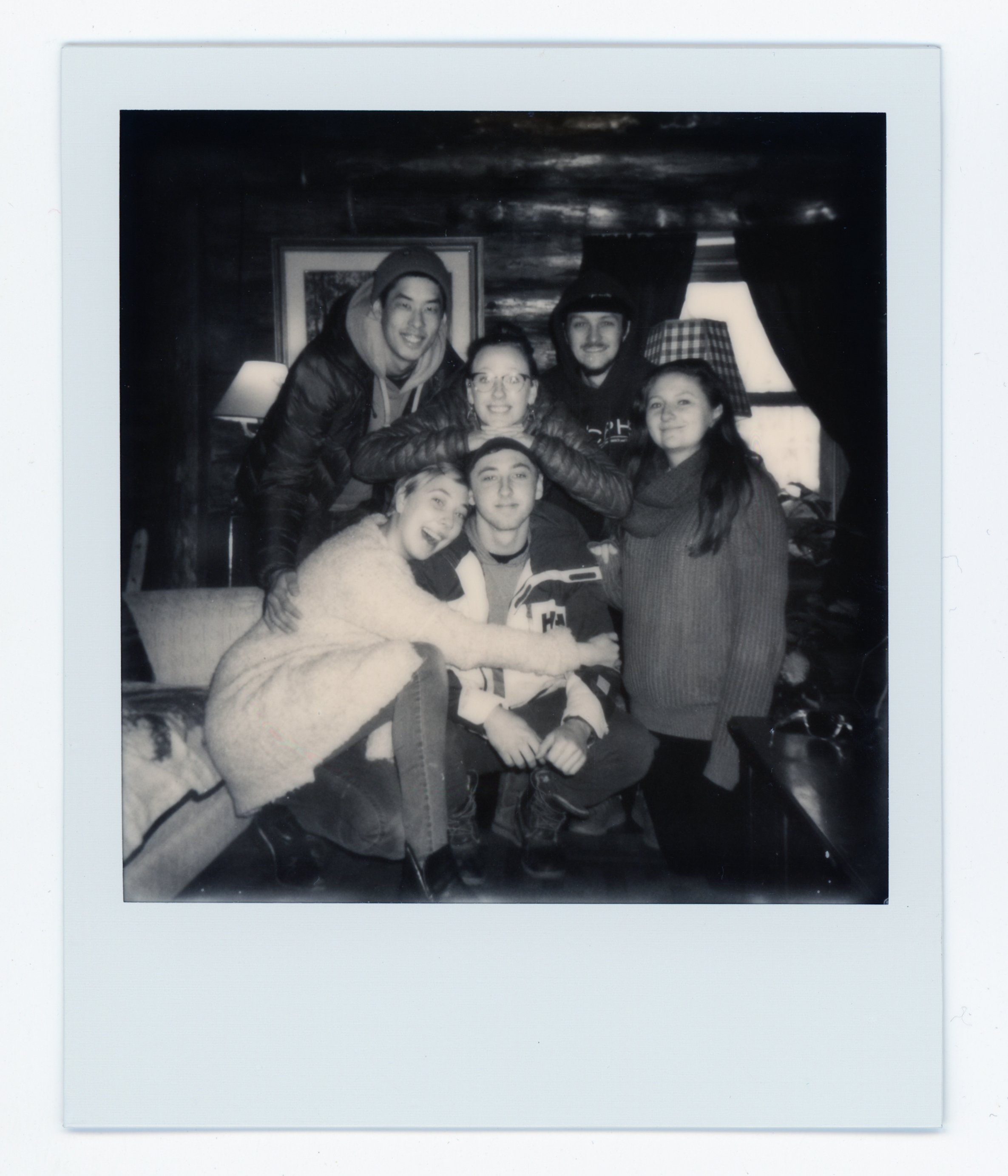 Black-and-white photograph of six young people smiling and gathered together in a cozy indoor setting with wooden walls and a window.