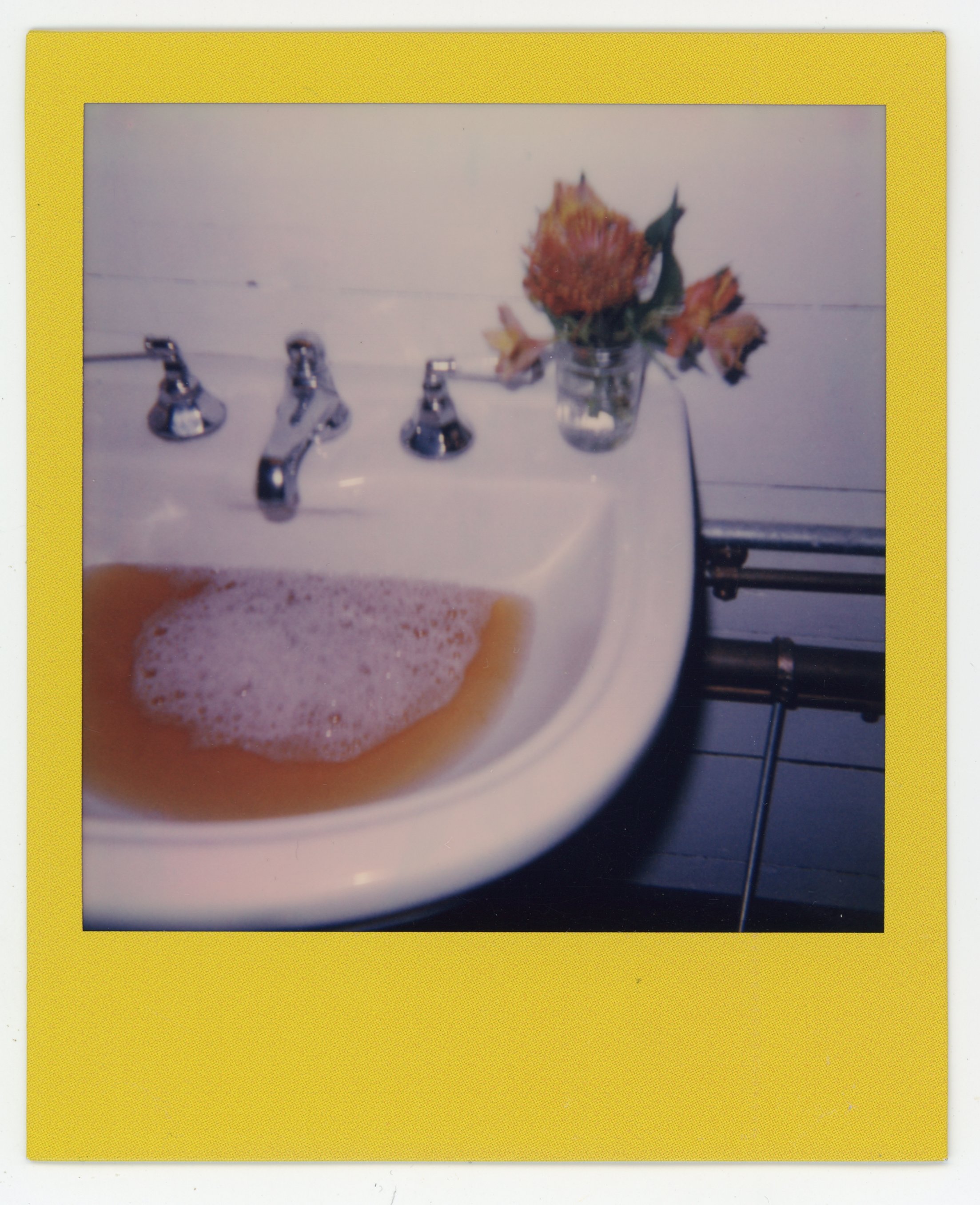 Vintage polaroid photo of a bathroom sink with soap bubbles, and a small bouquet of yellow and orange flowers in a glass jar placed on a white countertop.