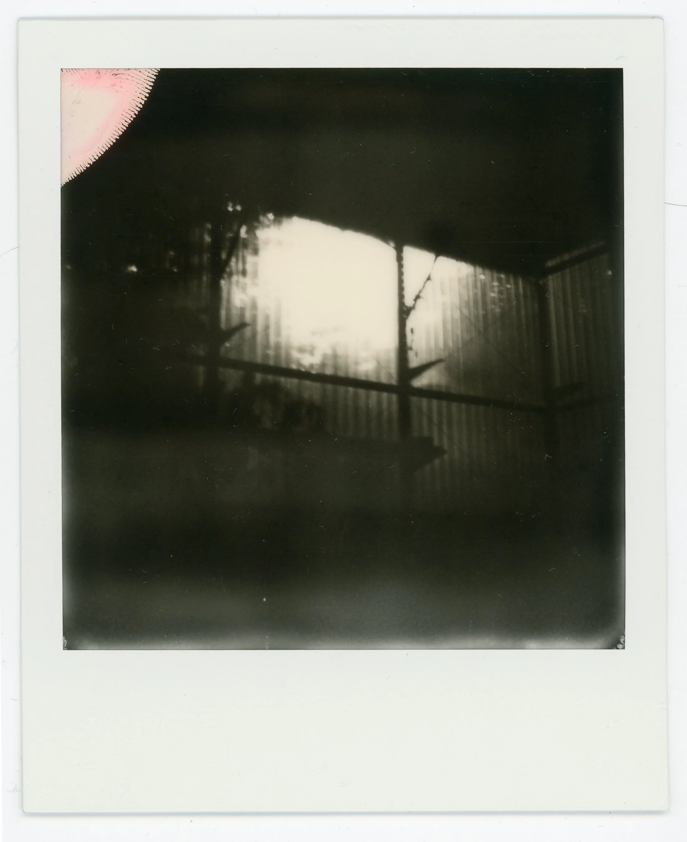 Black and white Polaroid photo of a porch with a railing, a corrugated metal wall, and a bright window or opening in the background, partially obscured by shadows.