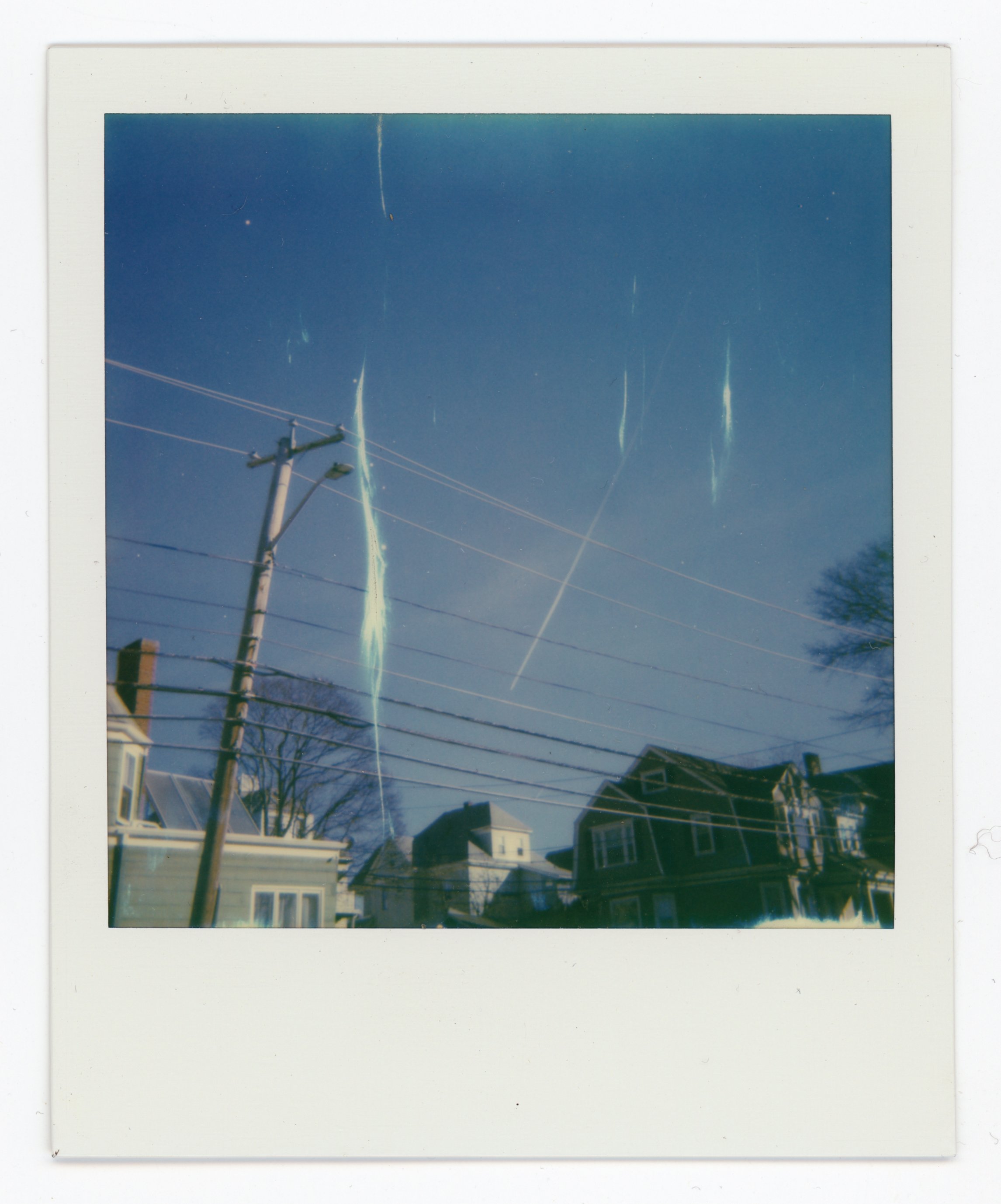 Polaroid photo of a clear blue sky with several streaks of light, some crossing, above residential houses and power lines.