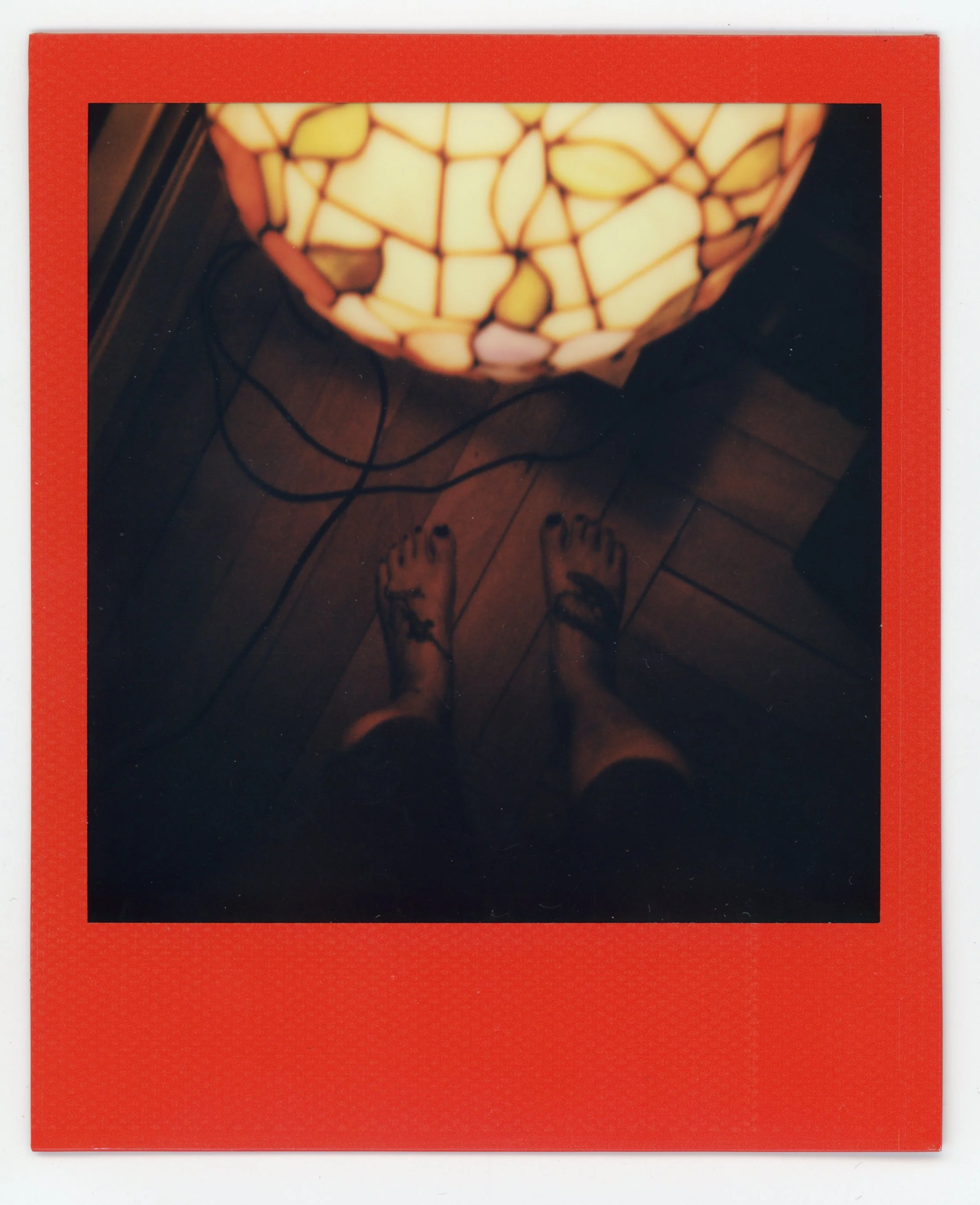 Photograph of a person’s feet with henna tattoos, standing on wooden floorboards, with a glowing mosaic lampshade in the upper part of the image, and power cords on the floor.