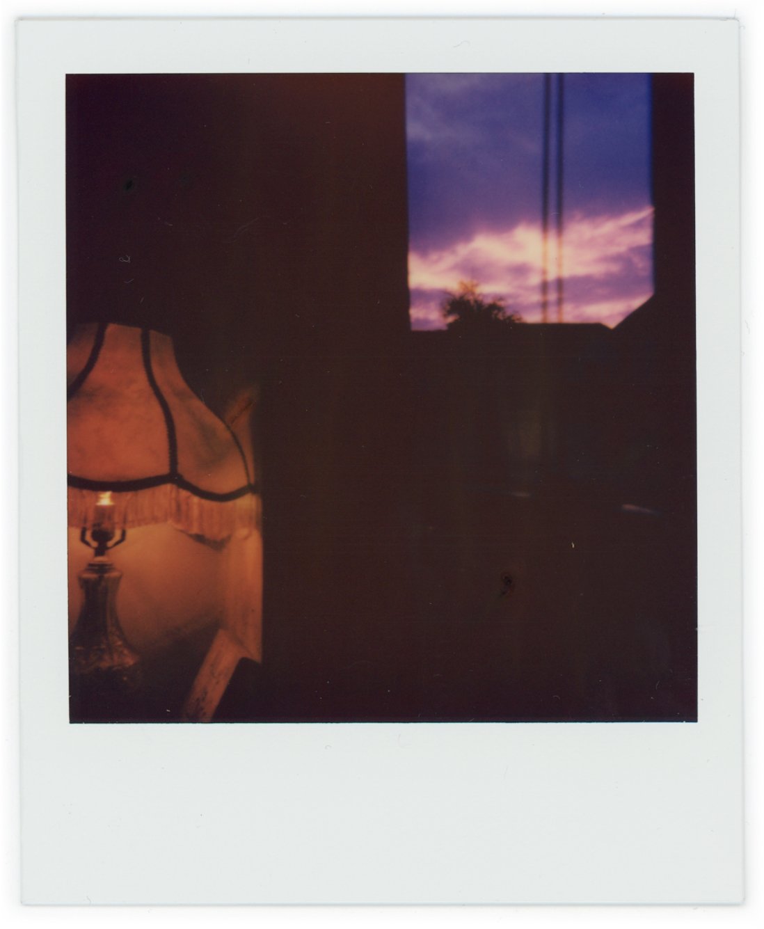 A silhouette of a person, part of a vintage lamp, and a window with a view of a colorful sunset sky with clouds and trees outside.