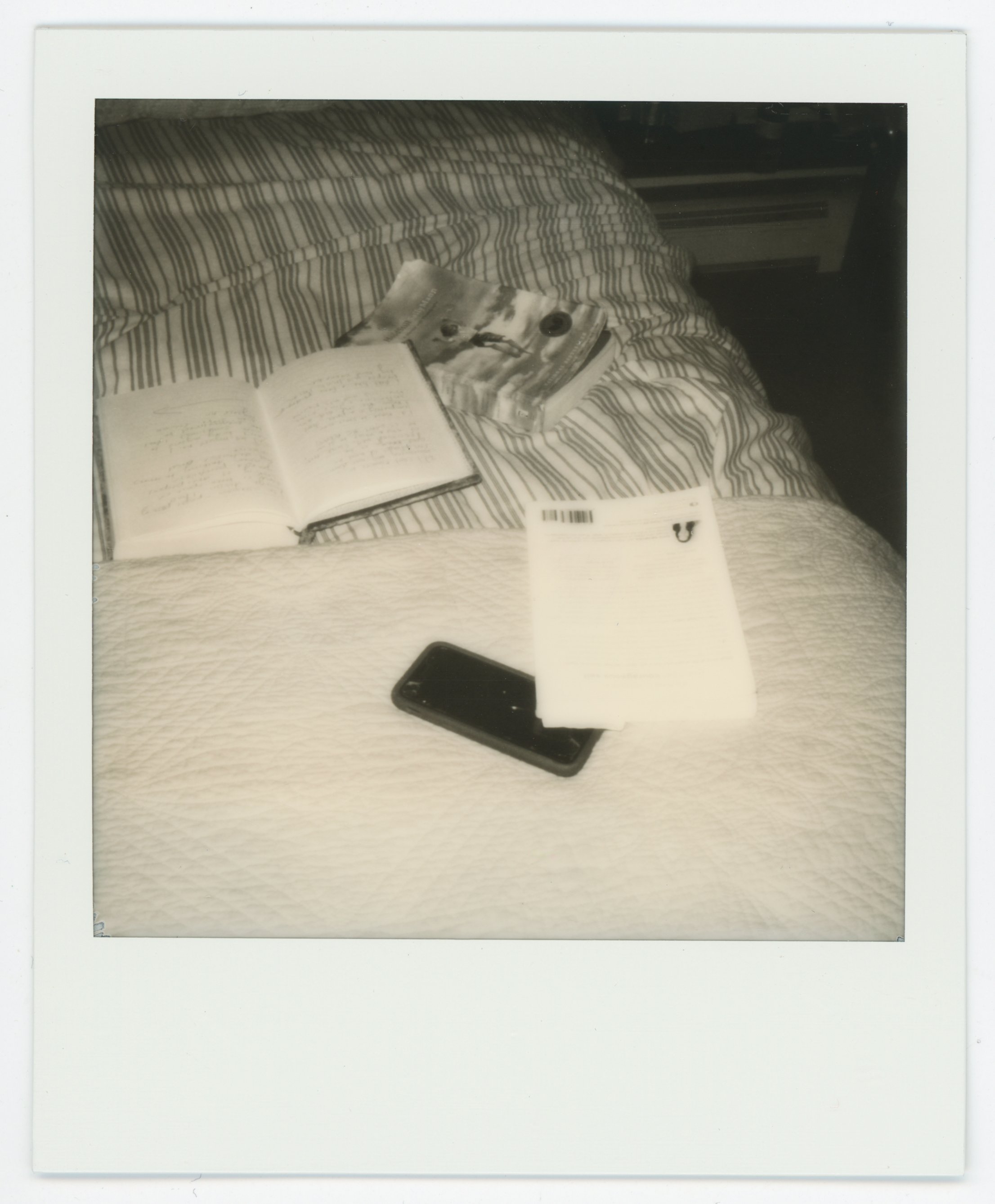 Polaroid photo of a bed with striped sheets, featuring an open notebook, a magazine, a smartphone, and a handwritten letter