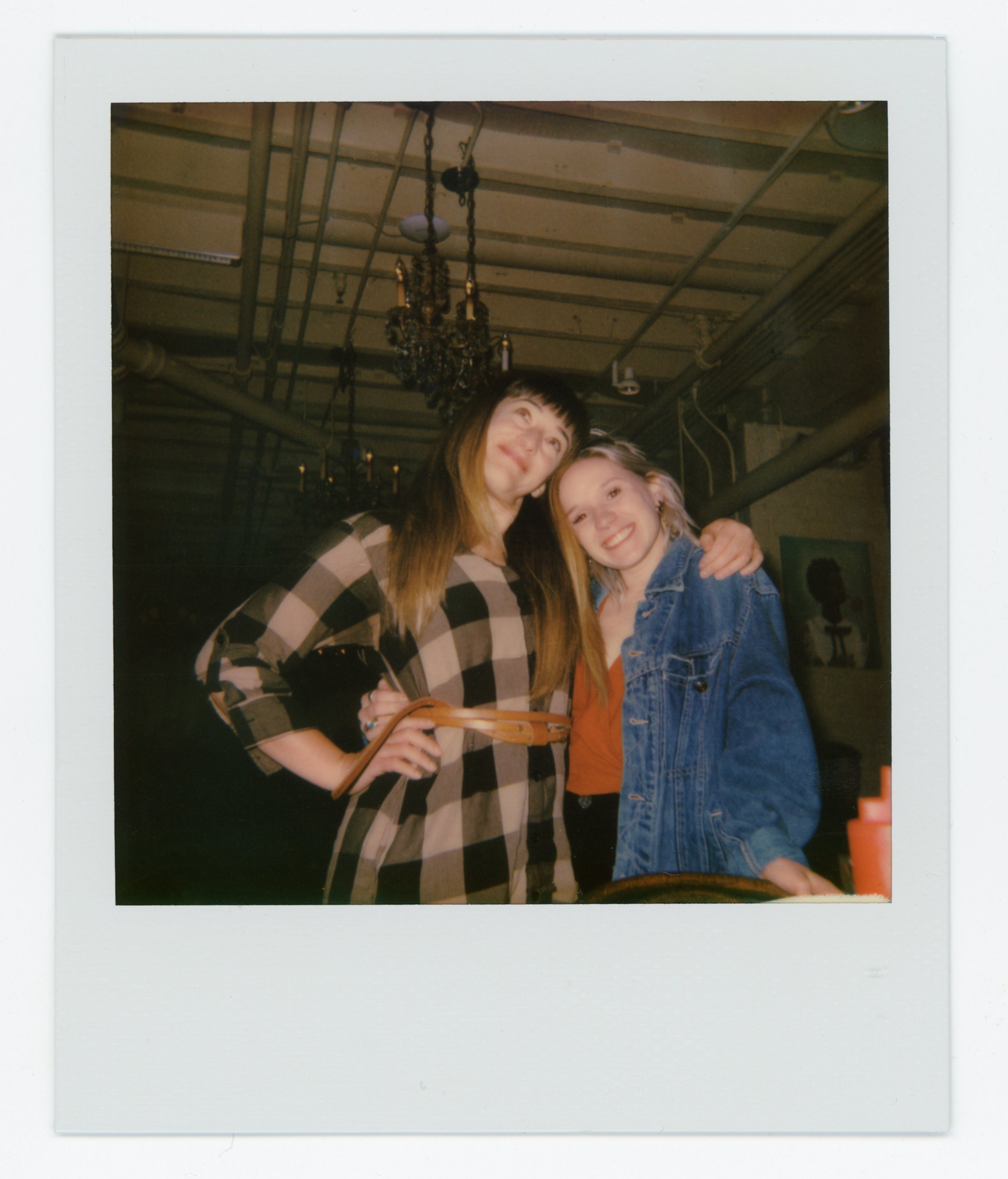 Two young women standing close together, smiling, in an indoor setting with chandeliers and industrial pipes in the background.