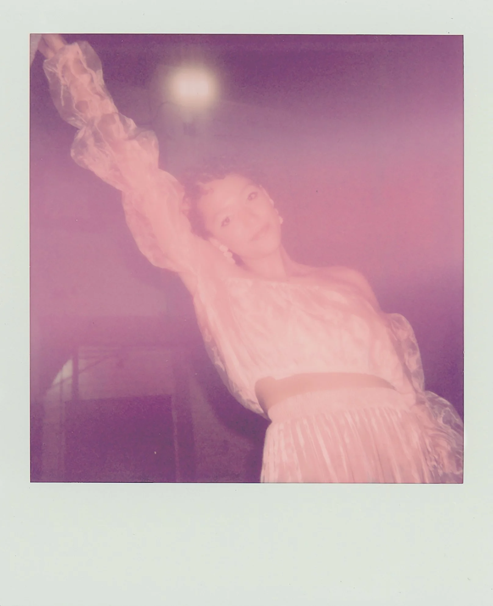 A woman in a pink dress with puffed sleeves, posing with her arm raised and a relaxed expression, in front of a dark background.