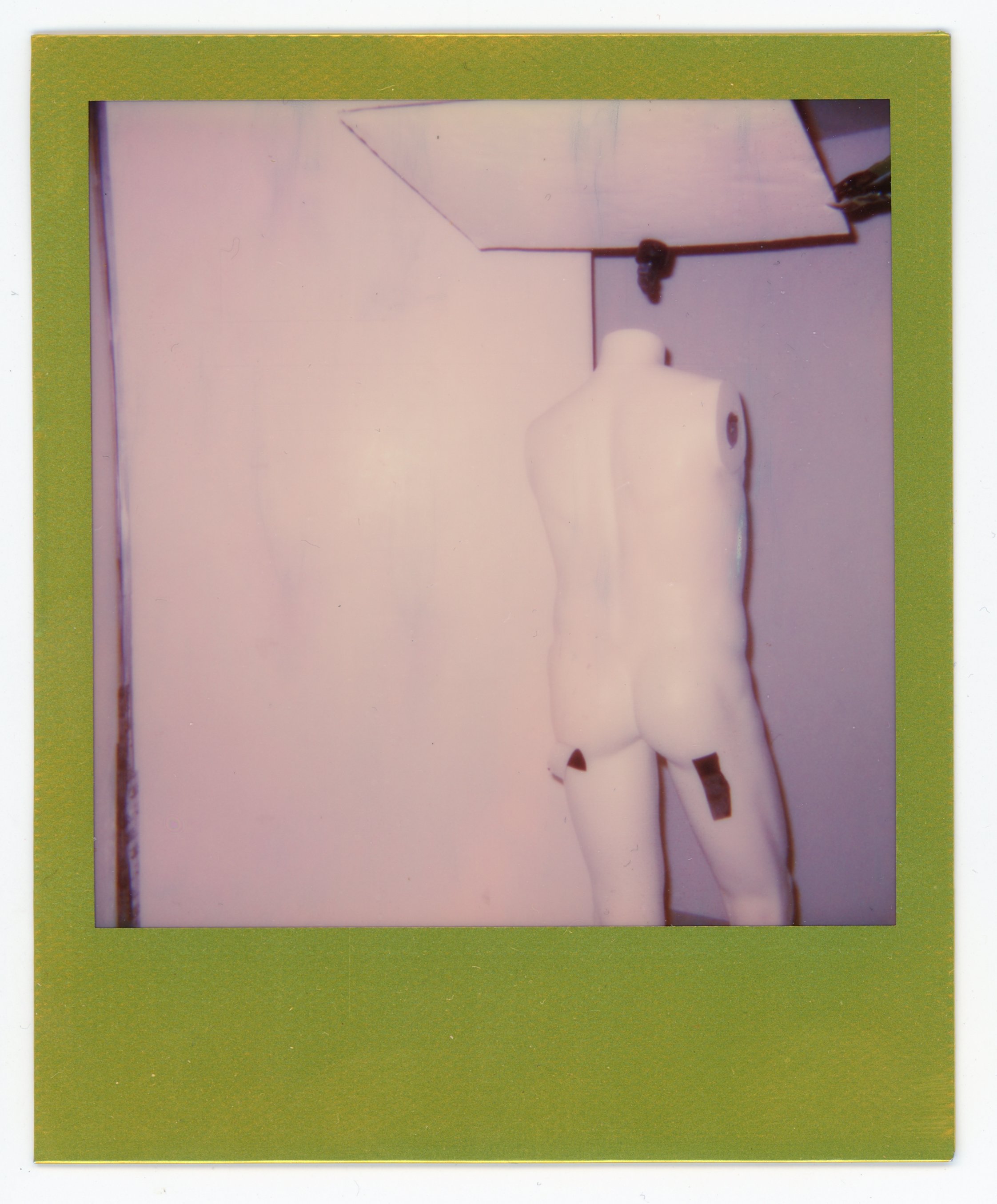 Polaroid photograph of a white mannequin torso with no head, arms, or legs, positioned against a wall, with a mirror in the background reflecting part of the scene.