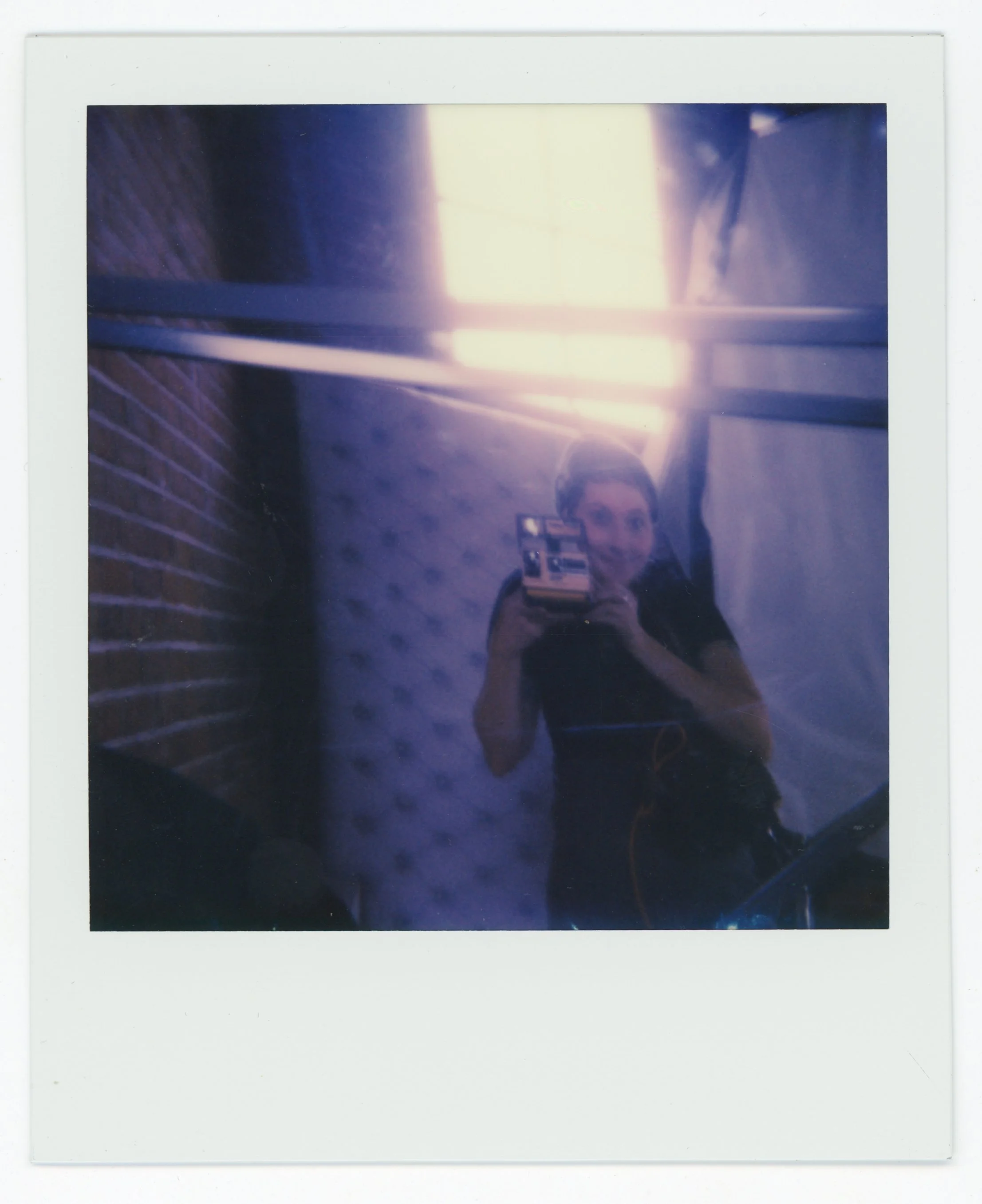 A woman taking a mirror selfie with a camera, visible in a dimly lit room with brick and fabric walls, and neon lighting overhead.