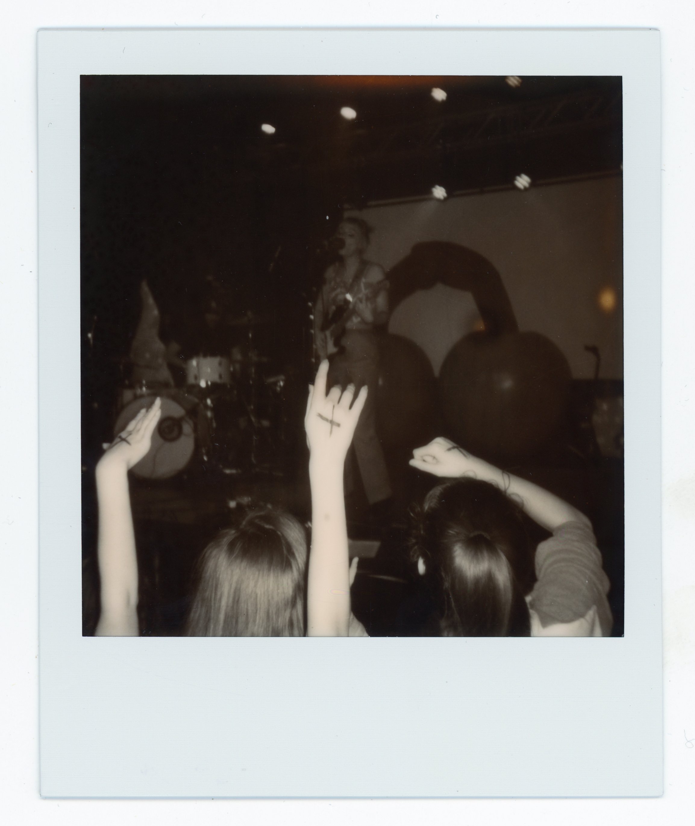 Black and white photo of a concert or performance with a woman singing on stage, silhouettes of large objects behind her, and audience members with hands raised in the foreground.