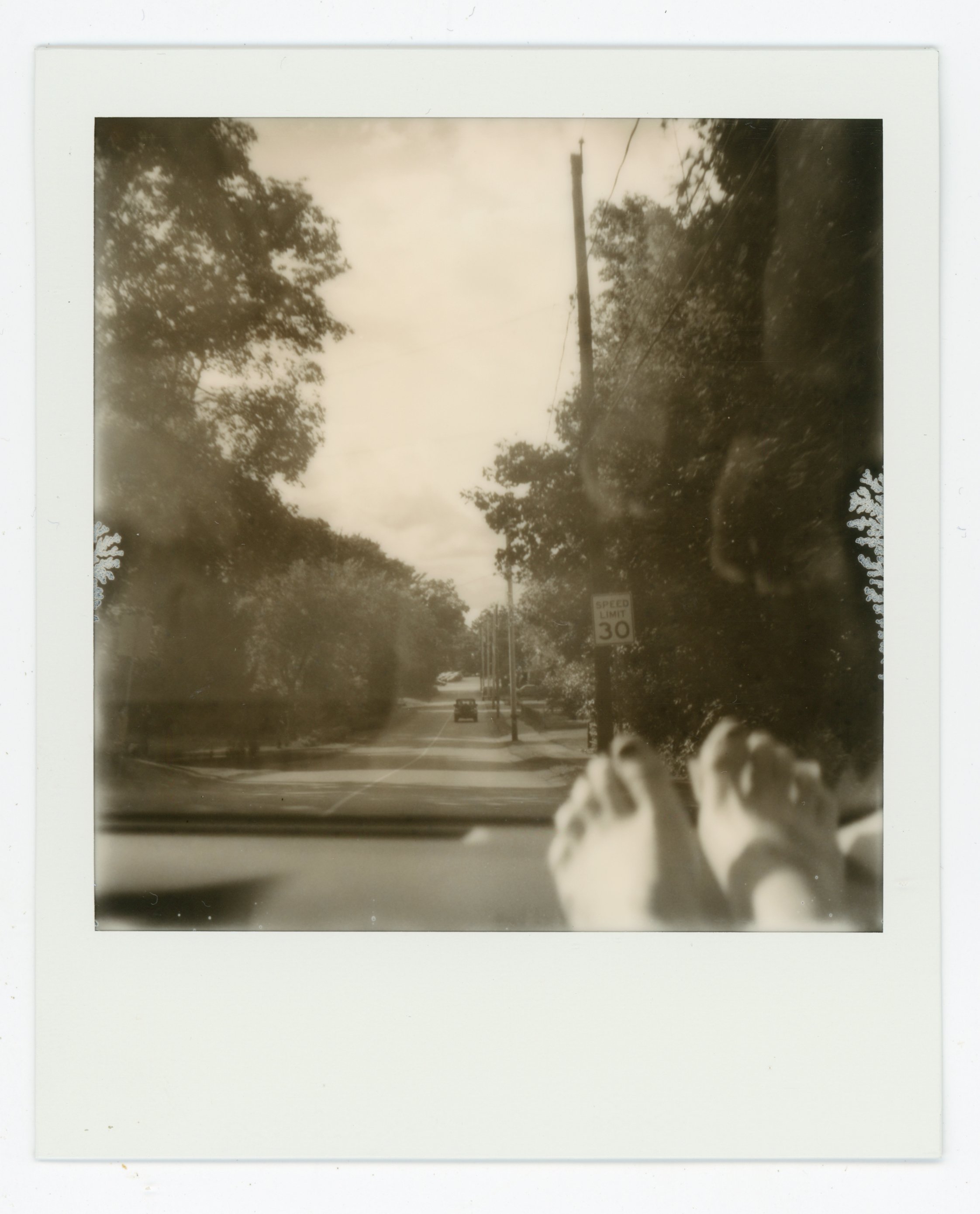 A black and white photograph taken from inside a car showing a view of a suburban street with trees, power lines, a speed limit sign of 30 miles per hour, and a car in the distance. The photographer's feet are visible in the foreground resting on the