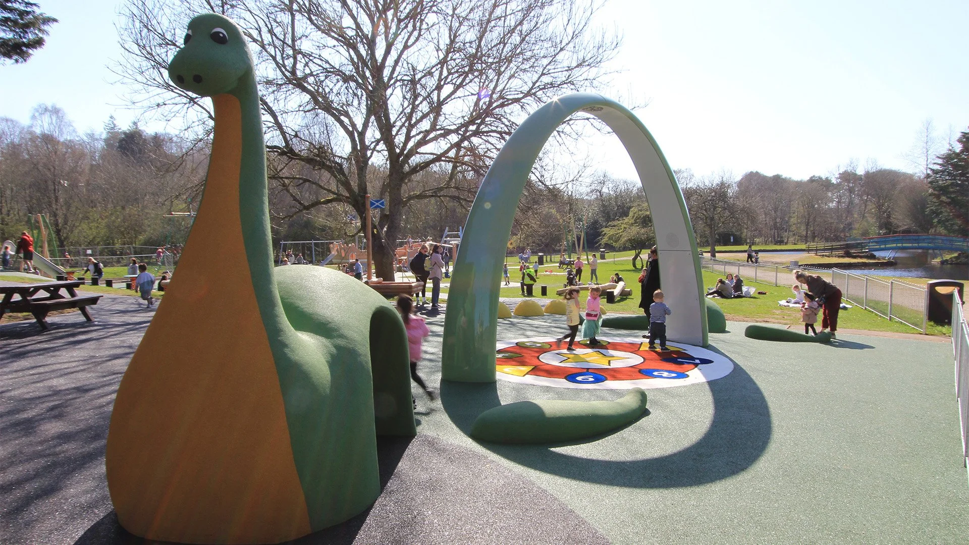 A playground with colourful fibreglass structures, including a Loch Ness Monster and a circular game area with children playing. There are adults supervising, trees in the background, and a pond nearby on a sunny day. Creative and Custom fibreglass.