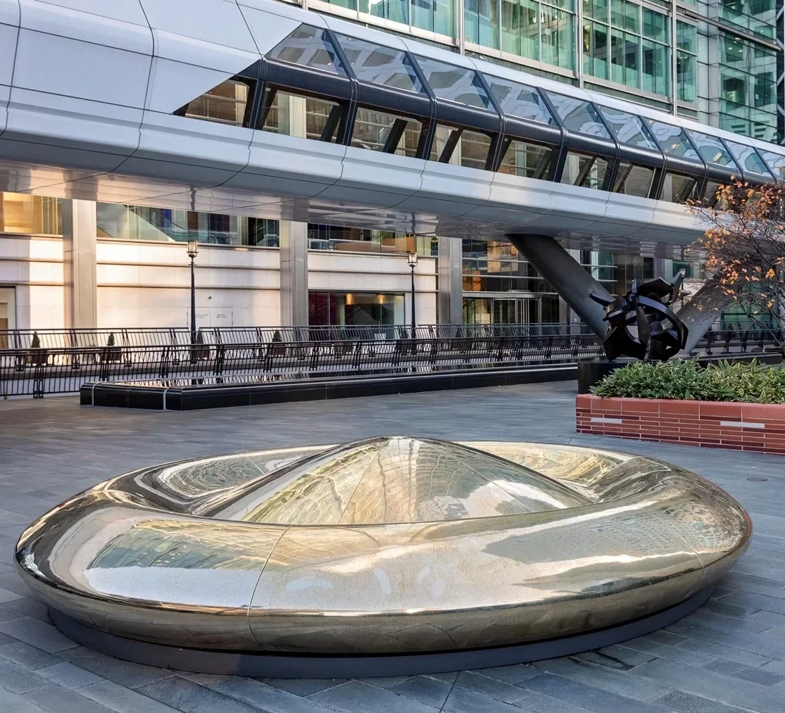 A modern urban scene featuring a sleek, metallic, boat-shaped bench or sculpture on a paved plaza, with a contemporary glass-covered bridge or walkway above and office buildings in the background. Sculpture. Art.  Creative and Custom fibreglass.